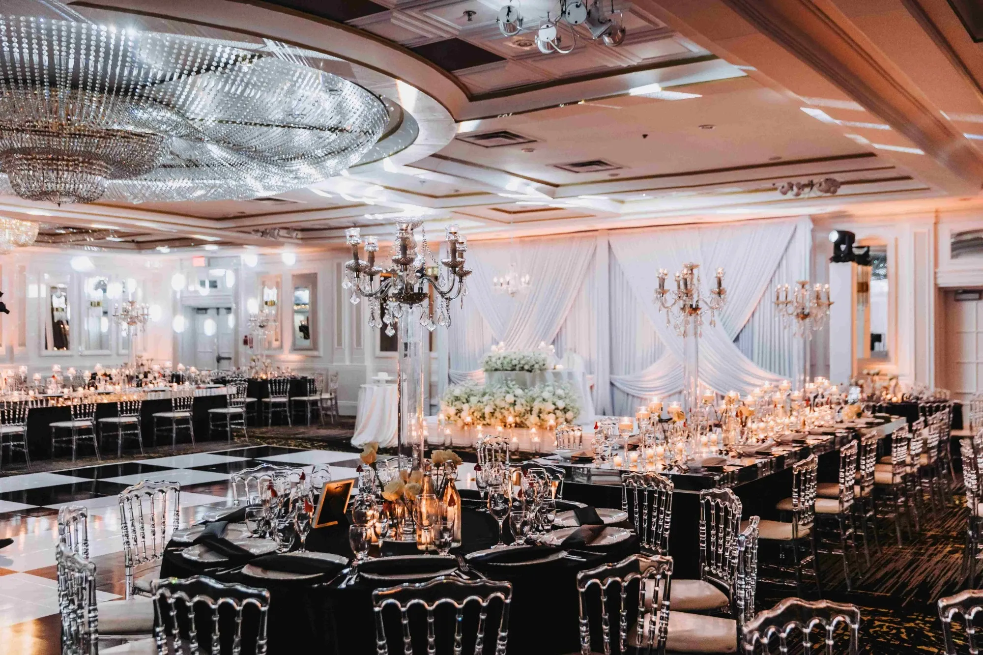 The image showcases a beautifully decorated wedding venue at Plaza Volare, categorized under Wedding Venues. The spacious room features an elegant setup with luxurious, crystal chandeliers hanging from the ceiling, casting a soft glow over the venue. Round tables are elegantly arranged, adorned with white tablecloths and intricate silver candelabras, creating a sophisticated ambiance. Each table is set with fine china, glassware, and floral centerpieces. The background features draped white fabric and a lush arrangement of white flowers, adding to the romantic atmosphere suitable for a wedding reception.