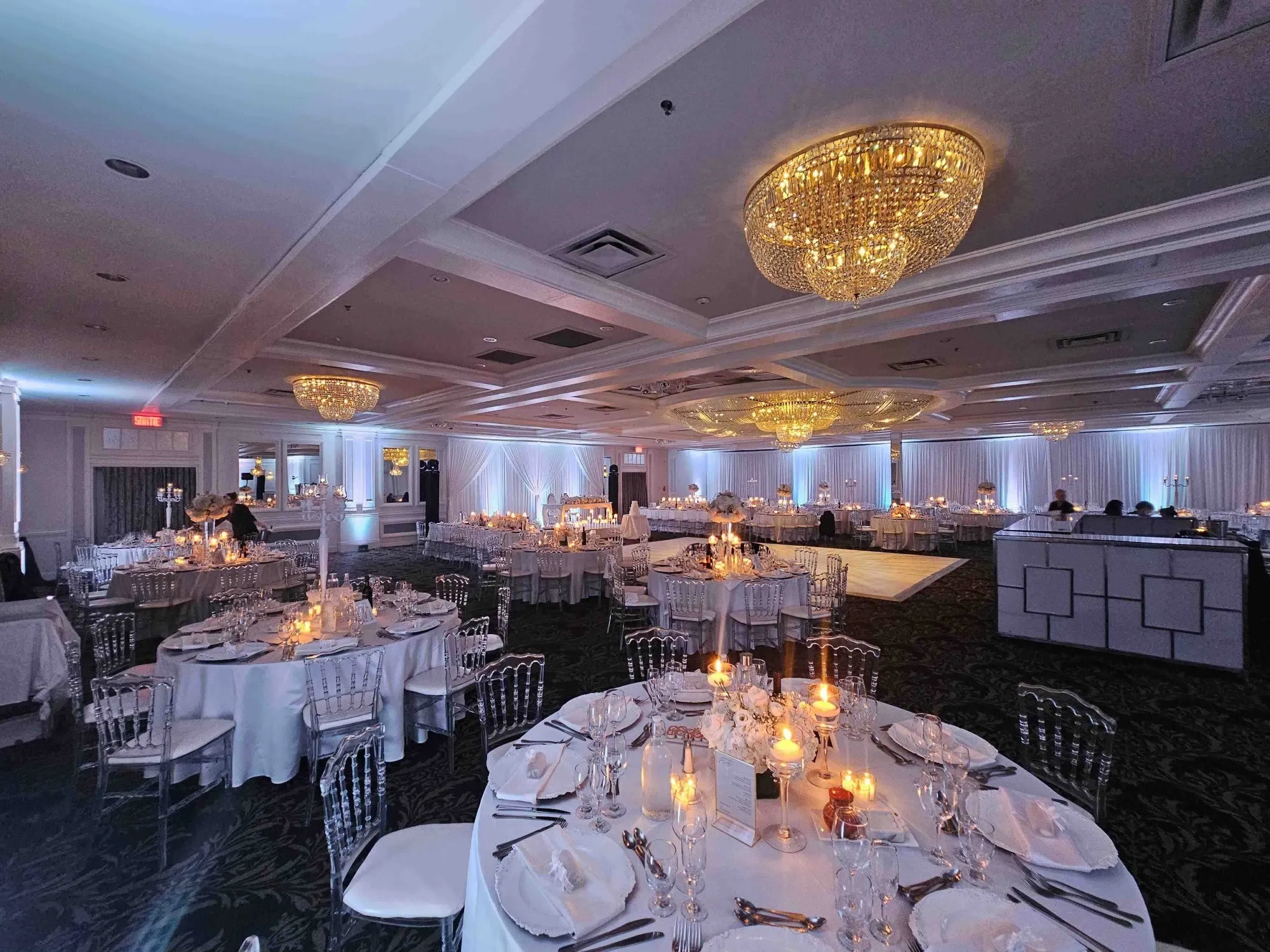 The image depicts a beautifully decorated wedding venue at Plaza Volare, a featured location in the Wedding Venues category on Mariage Elegant, Canada. The spacious hall is elegantly adorned with opulent chandeliers casting a warm glow over round tables set with pristine white tablecloths, fine china, and glassware. Each table is illuminated by soft candlelight, adding a romantic ambiance. The ceiling is lined with intricate molding, and a large dance floor is visible in the background, ready for guests to enjoy. Crystal-clear chairs complement the sophisticated setting, highlighting Plaza Volare’s attention to detail and luxurious atmosphere ideal for weddings.