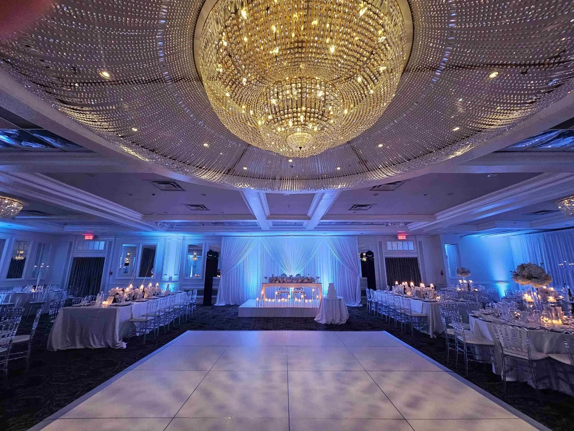 This image showcases the elegant interior of Plaza Volare, featured in the Wedding Venues category on Mariage Elegant, Canada. The spacious hall is beautifully decorated for a wedding reception, highlighted by a stunning, large crystal chandelier casting a warm glow across the room. The setup includes an expansive white dance floor at the center, surrounded by elegantly set tables adorned with white linens and flickering candles, creating a romantic ambiance. At the far end, a head table draped in white fabric is tastefully arranged, enhancing the sophisticated atmosphere suitable for a memorable wedding celebration.