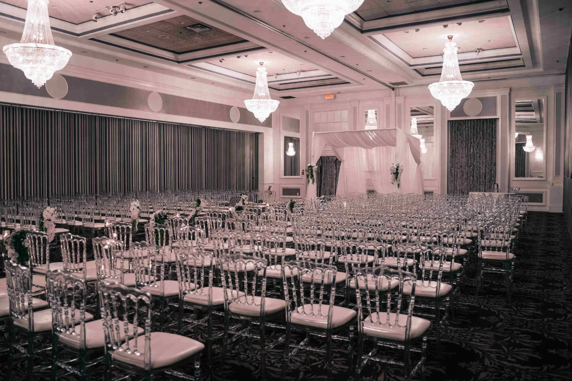 The image depicts the interior of Plaza Volare, a wedding venue listed on Mariage Elegant in Canada. The spacious room is elegantly set up with rows of transparent acrylic chairs arranged neatly, facing a central aisle. Overhead, large crystal chandeliers illuminate the space with a warm glow, complementing the sophisticated ambiance. In the background, there is a beautifully draped arch, suggesting a setup for a wedding ceremony. The walls are adorned with intricate paneling, enhancing the venue’s luxurious and refined appearance, making it an ideal location for hosting elegant weddings.