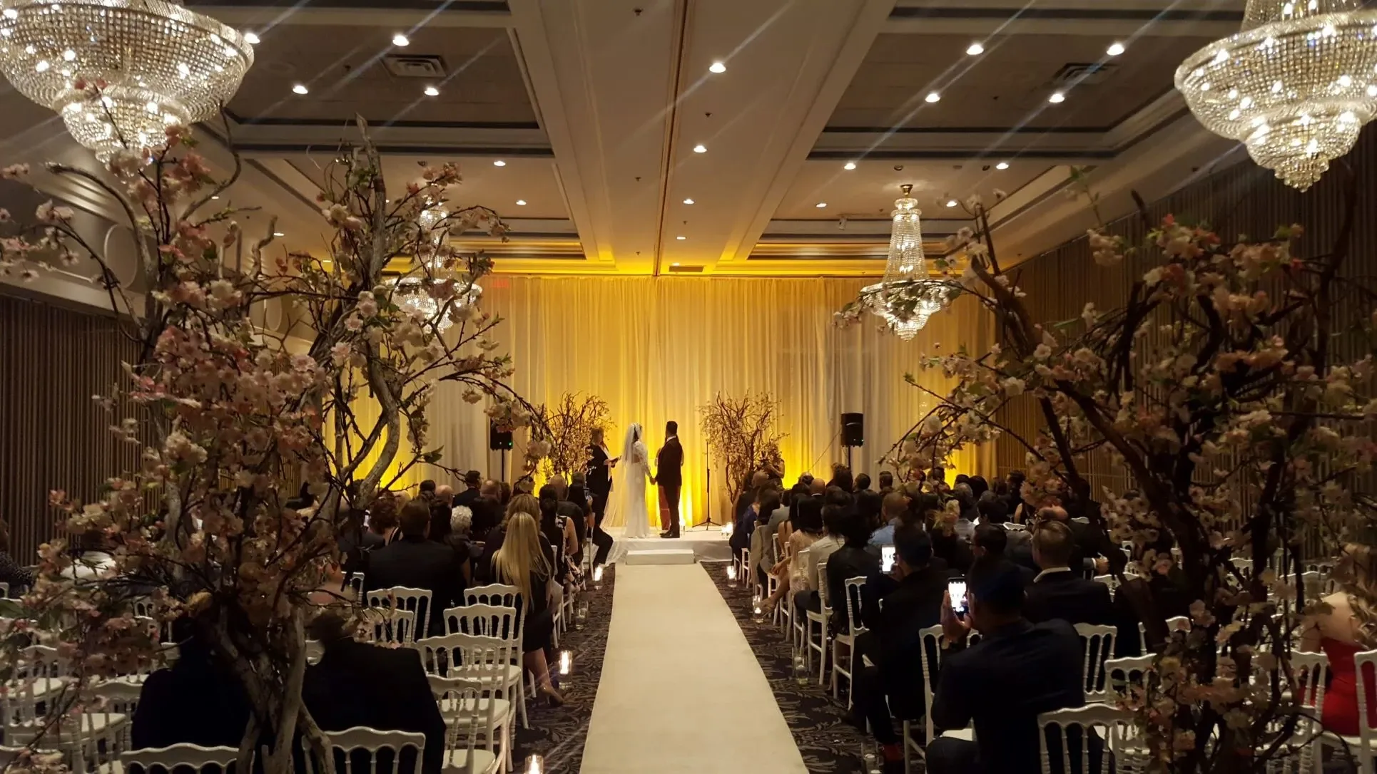 The image showcases a beautiful wedding ceremony held at Plaza Volare, a distinguished wedding venue. The elegant indoor setting features a long, white aisle leading to a softly lit altar adorned with yellow drapery. On either side of the aisle, guests are seated on white chairs, attentively watching the ceremony. The room is elegantly decorated with tall, blossoming tree branches flanking the aisle, and grand chandeliers hanging from the ceiling, contributing to the venue's sophisticated ambiance.