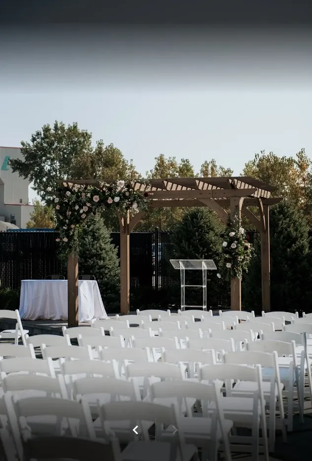 The image shows an outdoor wedding venue setup at Plaza Volare, categorized under Wedding Venues. There is a beautifully adorned wooden pergola surrounded by lush greenery, with elegant floral arrangements featuring white and pink flowers. In front of the pergola, rows of white chairs are neatly arranged for guest seating. A white table and a clear podium are also visible, suggesting a ceremonial setup. The natural backdrop and clear sky create a serene and inviting atmosphere, ideal for a wedding celebration.