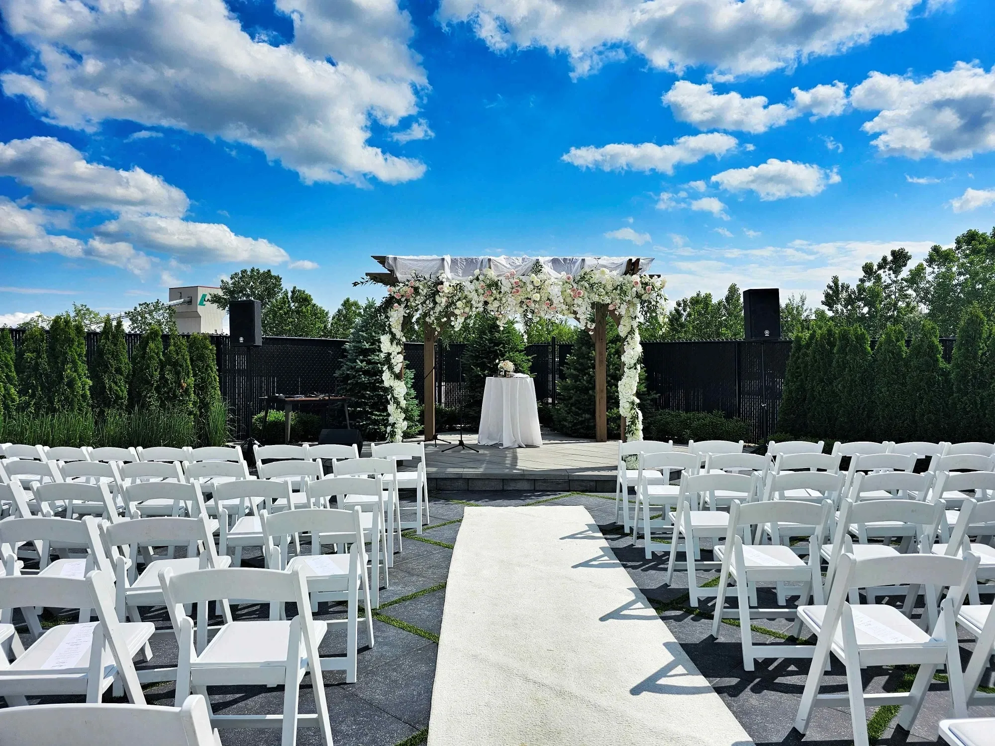 In this image of a wedding venue by Plaza Volare, part of the Mariage Elegant vendor listing in Canada, a beautifully arranged outdoor ceremony area is showcased. White chairs are neatly aligned on either side of an aisle, which is elegantly covered with a white runner. At the center, an arbor is adorned with lush white and pink floral arrangements, creating a stunning focal point. The ceremony space is surrounded by manicured greenery and trees, offering a serene and picturesque setting under a clear blue sky with scattered fluffy clouds. This setup highlights Plaza Volare’s dedication to creating memorable wedding experiences.