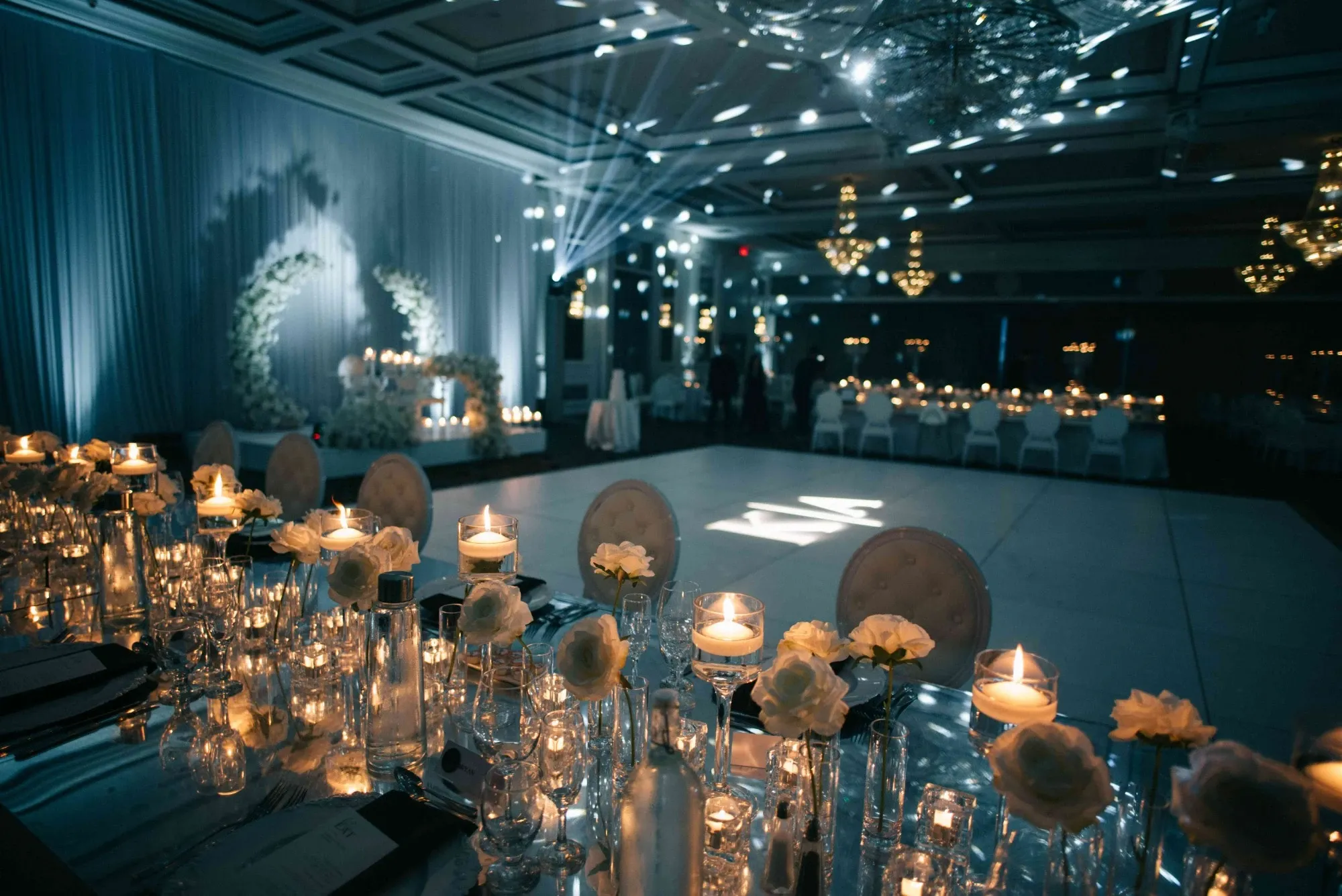 The image showcases an elegant wedding venue by Plaza Volare, categorized under Wedding Venues, at Mariage Elegant in Canada. The ballroom is beautifully decorated with a polished dance floor centrally located under soft, ambient lighting. White floral arrangements and candles line the tables, adding a romantic and luxurious touch to the setting. The ceiling features intricate designs and glowing chandeliers, enhancing the sophisticated atmosphere perfect for a wedding celebration.