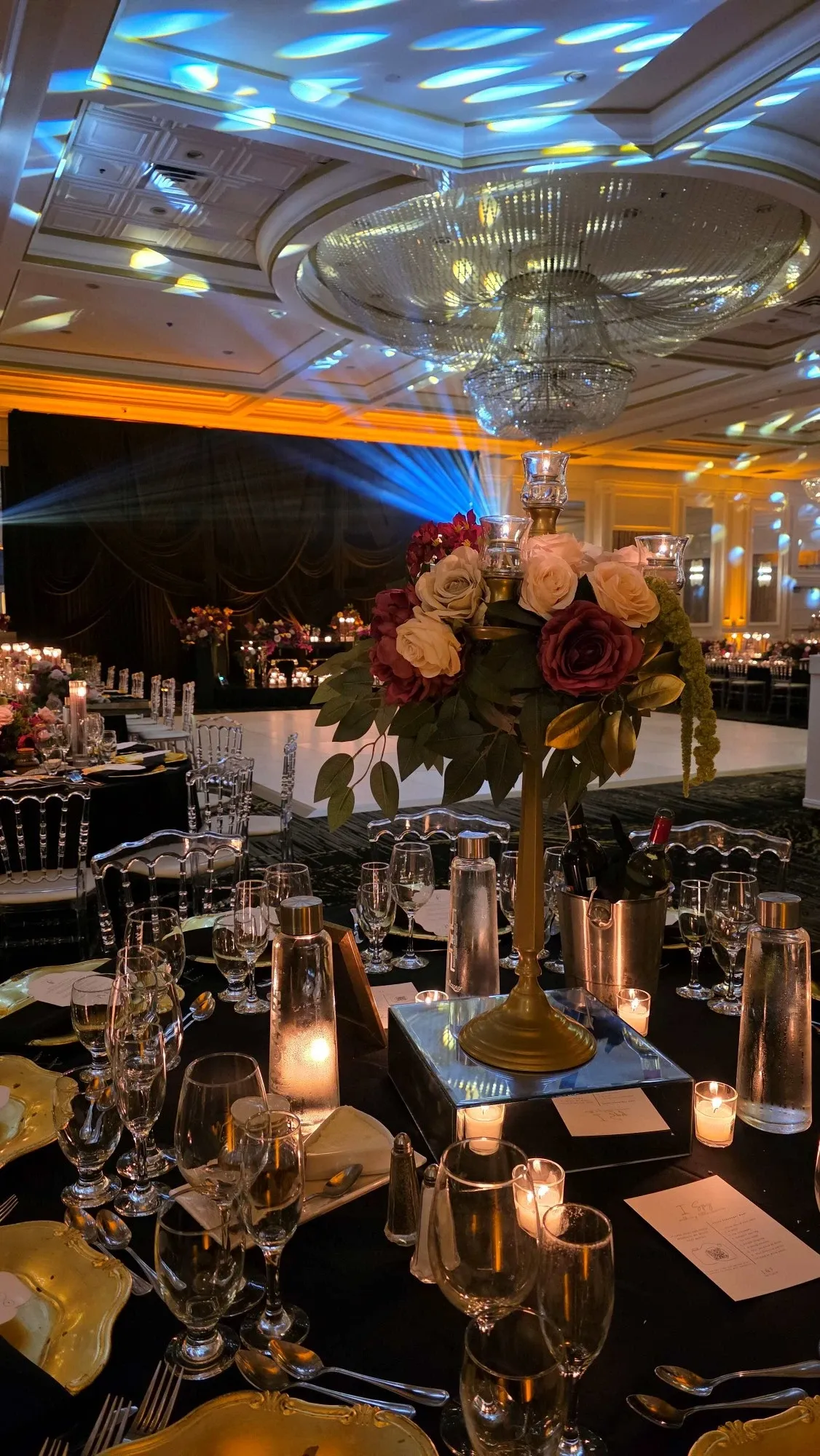 The image showcases an elegantly decorated wedding venue at Plaza Volare, highlighting its suitability as a prime wedding location. The setting features a beautifully arranged table centerpiece of roses in shades of burgundy and cream with lush greenery, situated on a stylish stand. Surrounding the floral arrangement are intricately set tables, complete with fine glassware and neatly folded napkins, all illuminated by the warm glow of candlelight. The ambient lighting casts a golden hue across the room, enhancing the upscale atmosphere. A large, ornate chandelier hangs from the ceiling, adding sophistication to the venue. This image captures the grandeur and refined ambiance that Plaza Volare offers in its wedding venue category.