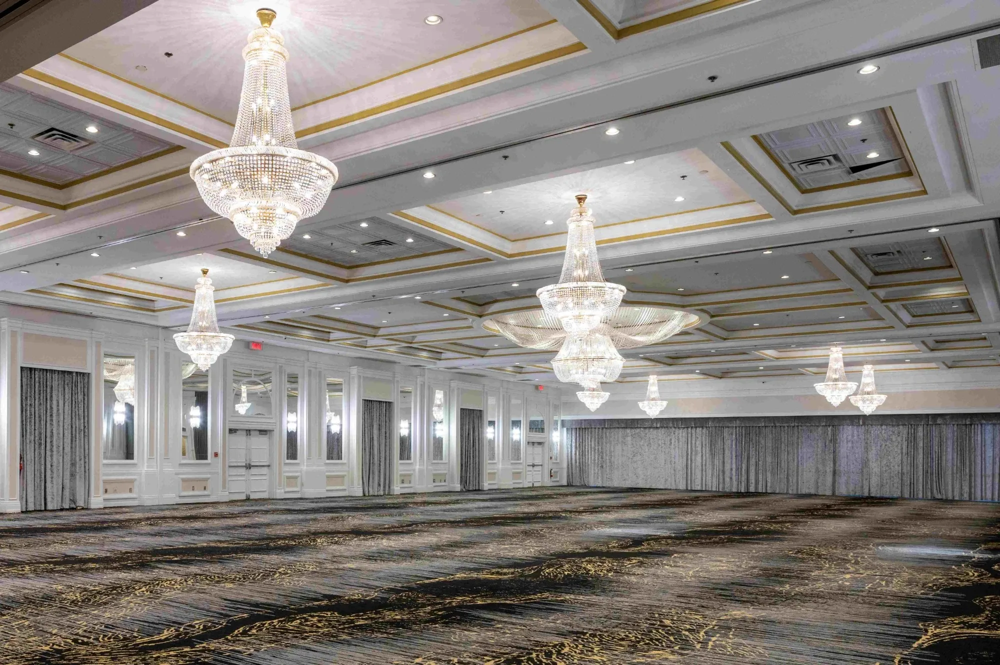 The image showcases an elegant and spacious wedding venue by Plaza Volare, categorized under Wedding Venues. The grand ballroom features luxurious crystal chandeliers hanging from a high ceiling with gold accents, creating a sophisticated ambiance. The room's walls are adorned with tall mirrors and decorative paneling, enhancing the sense of space and style. The floor is covered with a plush, patterned carpet that complements the elegant decor. This setting, perfect for a wedding reception or ceremony, offers a timeless and refined atmosphere for special occasions.