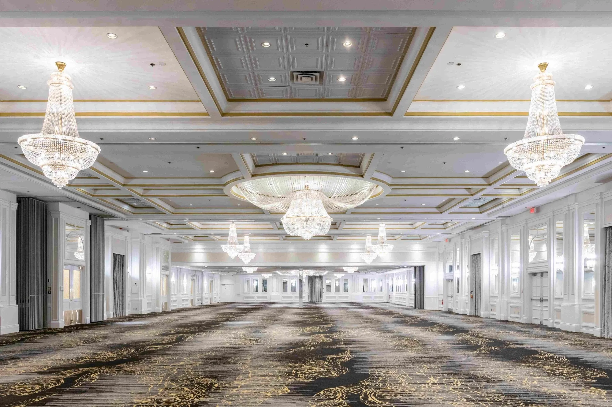 The image showcases Plaza Volare, a premier wedding venue in Canada, listed under the wedding planning and vendor category at Mariage Elegant. The spacious interior features an elegant ballroom with luxurious decor. The ceiling is adorned with several large, sparkling crystal chandeliers, casting a sophisticated glow across the room. The ballroom's ceiling is intricately designed with recessed lighting and coffered details, adding to the grandeur of the space. The carpet displays an intricate pattern in neutral tones, complementing the overall sophisticated ambiance. Tall, mirrored wall panels enhance the room's spaciousness, making it an ideal setting for a lavish wedding celebration.