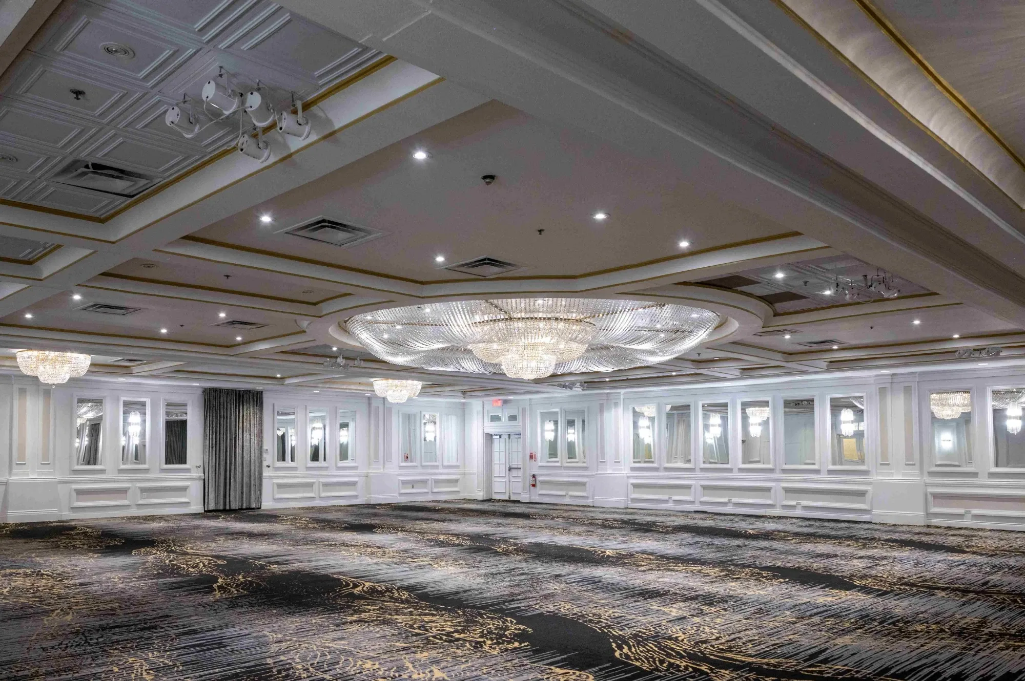 The image features Plaza Volare, a premier wedding venue in Canada, showcasing an elegant and spacious ballroom. The room is adorned with a large, central crystal chandelier surrounded by smaller chandeliers, providing a luxurious ambiance. The ceiling is intricately designed with coffered panels and gold accents, adding to the sophisticated décor. The walls are decorated with ornate moldings and large mirrors, enhancing the sense of space and opulence. The carpet features a contemporary pattern, complementing the room's grandeur, making it an ideal setting for a memorable wedding event.