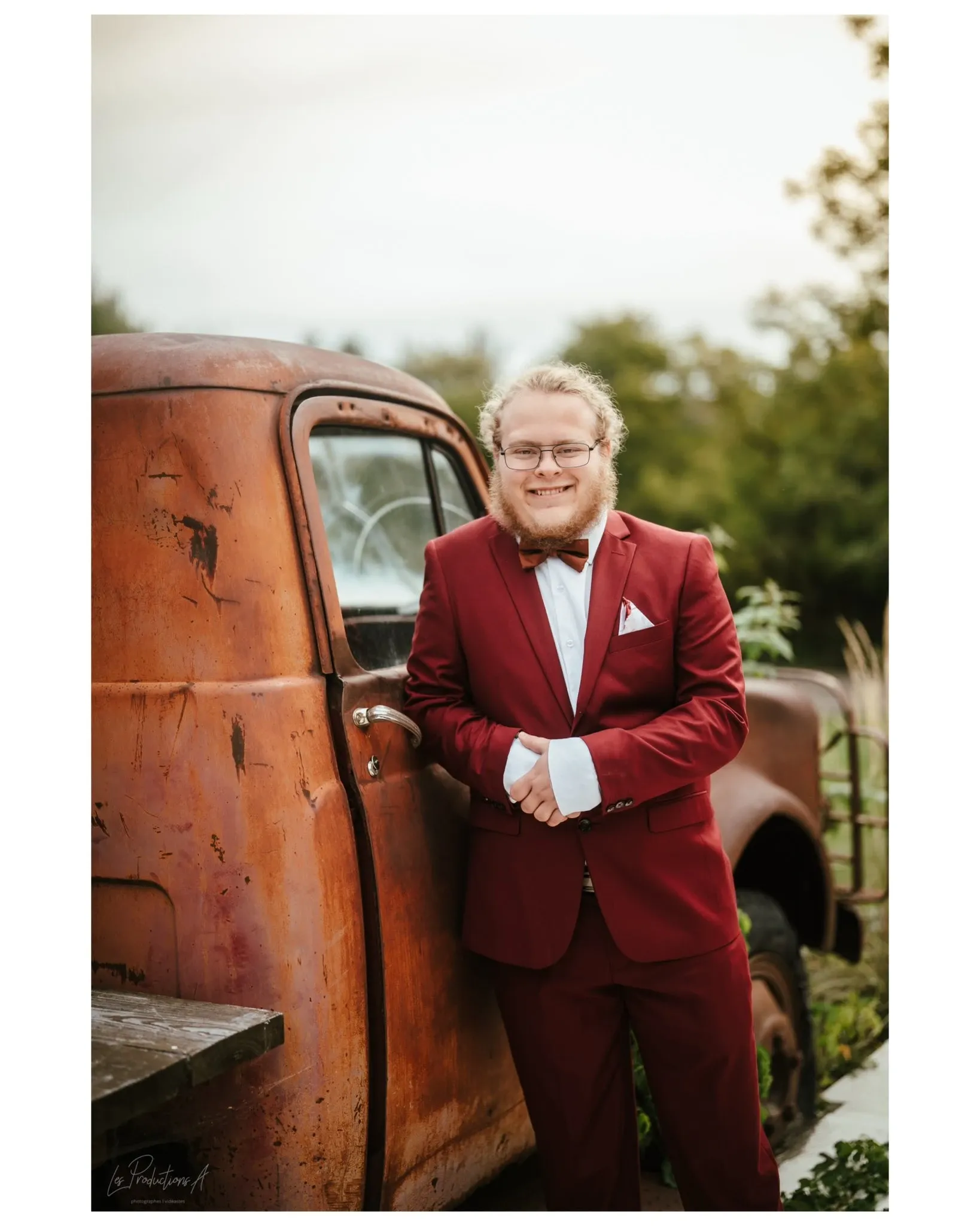 This image features a person dressed in a stylish maroon suit with a white shirt and bow tie, leaning against a rustic, vintage truck with a weathered appearance. The background includes greenery, indicating an outdoor setting, possibly a garden or rural venue, which adds a charming and timeless feel to the scene. The serene expression and relaxed pose convey a sense of calm and elegance, ideal for capturing memorable wedding moments. This image represents the work of Les Productions A, a vendor specializing in wedding photography, as featured on Mariage Elegant, Canada.