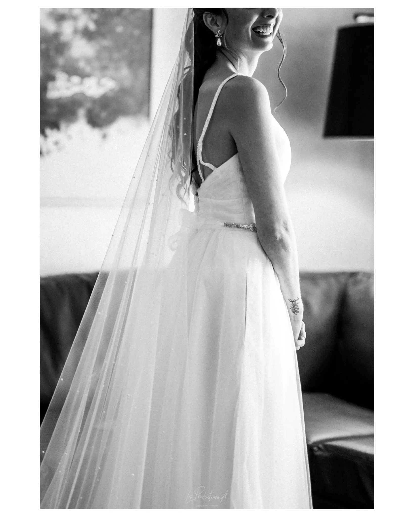 A black and white photograph by Les Productions A, showcasing a bride in an elegant wedding dress, highlighting the skilled wedding photography services available in Canada. The bride is standing indoors with a soft, natural expression, wearing a classic gown with a beautifully flowing veil. Her dress is complemented by a delicate belt, and she has graceful curls cascading from her elegant updo. The background features a blurred painting and a lamp, adding a cozy, intimate atmosphere to the image, perfectly capturing a special moment on her wedding day.