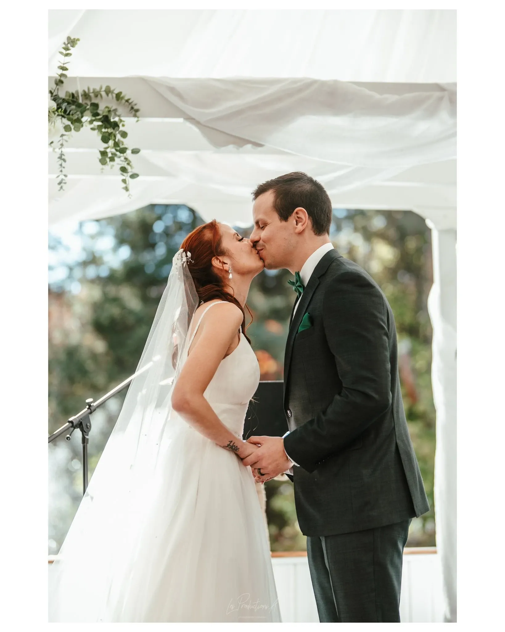 This image showcases a romantic moment captured by Les Productions A under the Wedding Photography category. A bride and groom share a kiss during their wedding ceremony, standing beneath a beautifully draped gazebo adorned with delicate greenery. The bride is wearing an elegant white gown with a veil, while the groom is dressed in a classic dark suit with a green pocket square. The natural light filters through the trees in the background, adding a serene and picturesque ambiance to this special occasion.