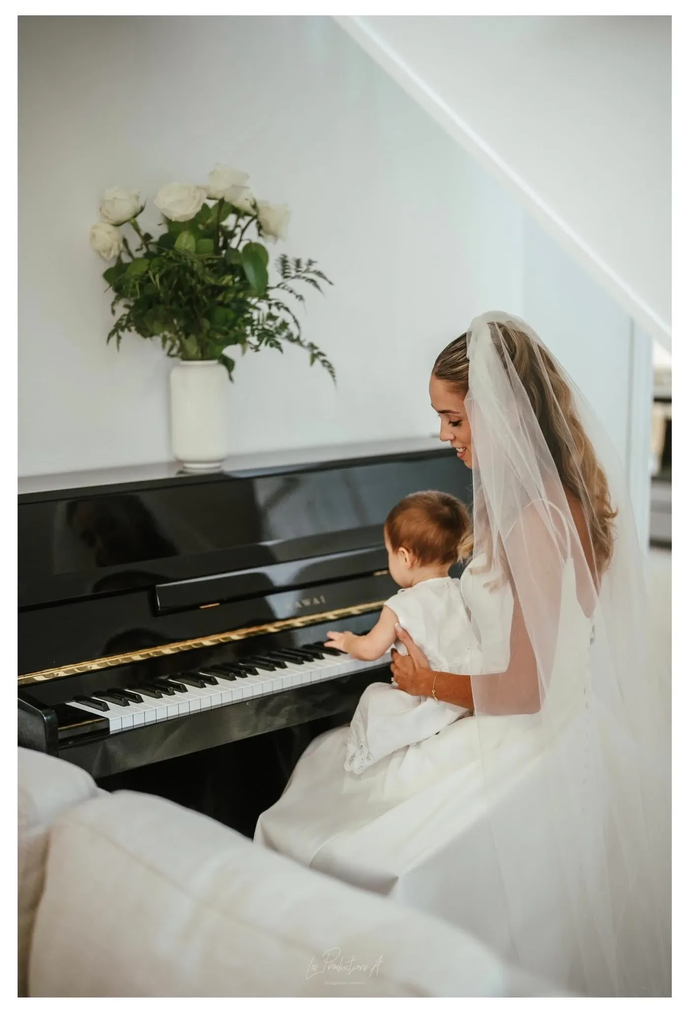 A bride in an elegant white gown and a long veil sits at a polished black piano, gently holding a small child dressed in a white outfit on her lap. Both are engaged in playing the piano keys softly. The serene scene is complemented by a vase of fresh white roses and greenery placed atop the piano, adding to the romantic atmosphere. This image showcases a tender moment likely captured during a wedding ceremony, part of the exceptional wedding photography services provided by Les Productions A, featured in the Wedding Photography category on Mariage Elegant, Canada.