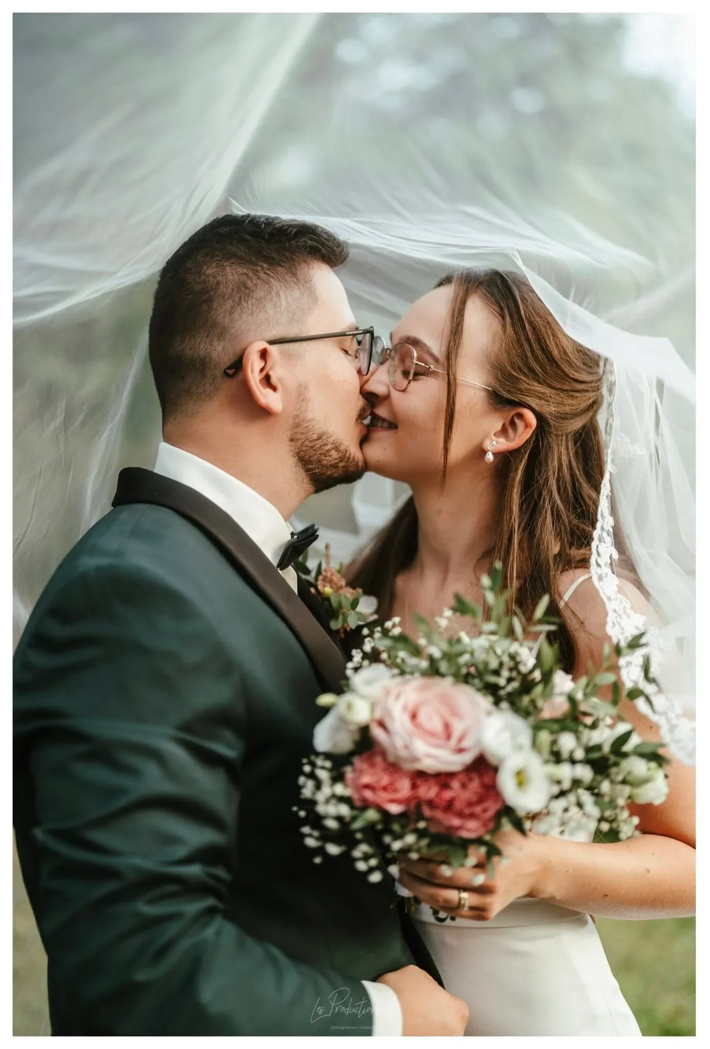 A bride and groom, elegantly dressed, share a joyful and intimate moment under a sheer wedding veil outdoors. The groom, in a dark suit and bow tie, leans in to kiss the bride, who is wearing a white gown and glasses. She holds a beautiful bouquet featuring pink and white flowers with greenery. The background is softly blurred, highlighting the couple’s happiness and love. This image showcases the expertise of Les Productions A in the Wedding Photography category, capturing special moments for Mariage Elegant in Canada.