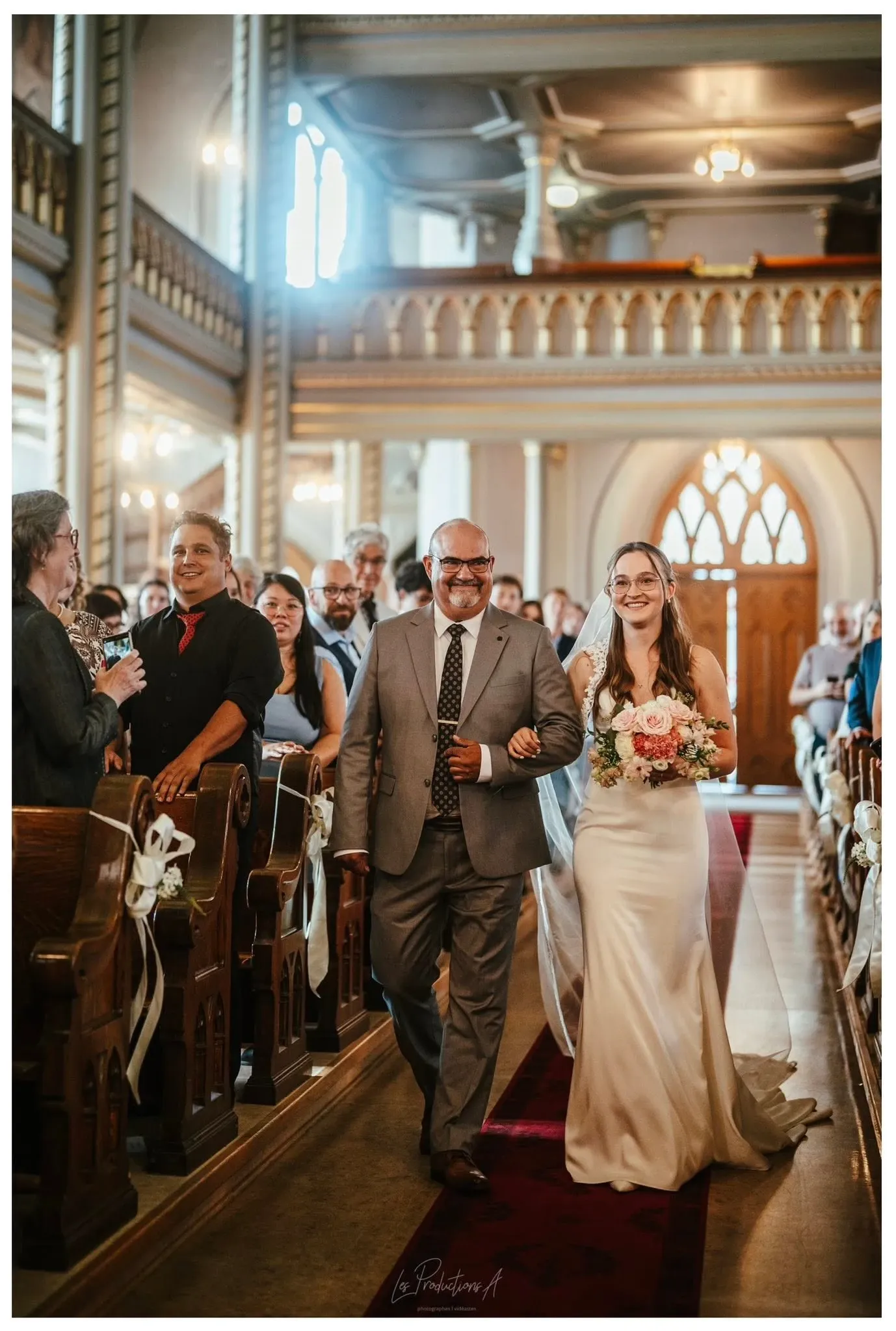 A bride in an elegant white gown walks down the aisle with a man, likely her father, in a beautifully decorated church. The aisle is lined with guests seated on wooden pews adorned with white ribbons, and the church's architecture features ornate railings and large arched windows. The bride smiles, holding a bouquet of pink and white flowers. Sunlight streams through the windows, creating a warm and inviting atmosphere. This image is a demonstration of the wedding photography services by Les Productions A, part of Mariage Elegant's vendor listings in the wedding category.