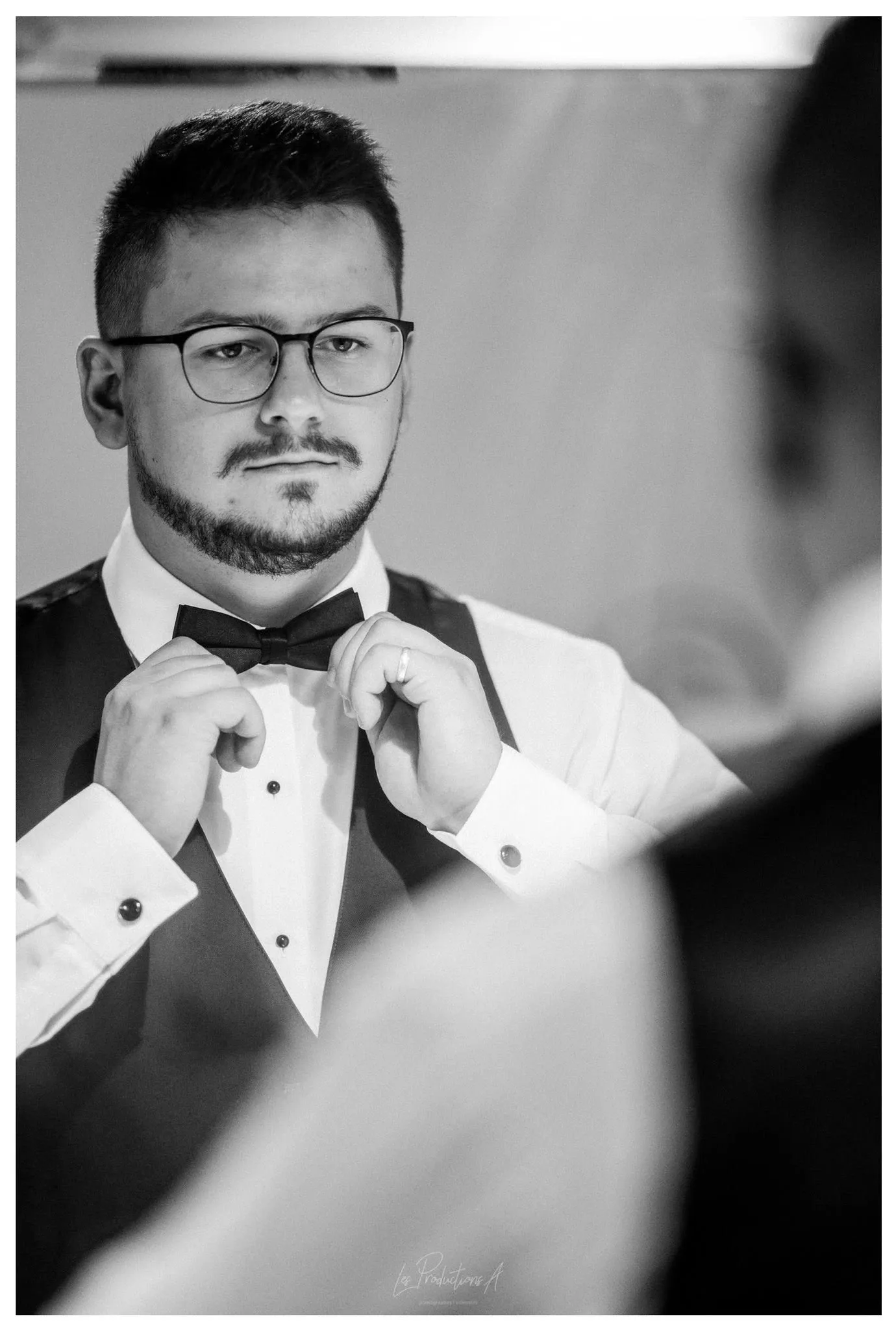 A groom is adjusting his bow tie while looking in a mirror, captured in a black and white photograph. He wears glasses and a formal suit with a vest, emphasizing a classic wedding look. This image highlights a moment of preparation and attention to detail, essential in the wedding photography category. The photograph, taken by Les Productions A, showcases the elegance and style important for weddings, featured on Mariage Elegant, Canada.