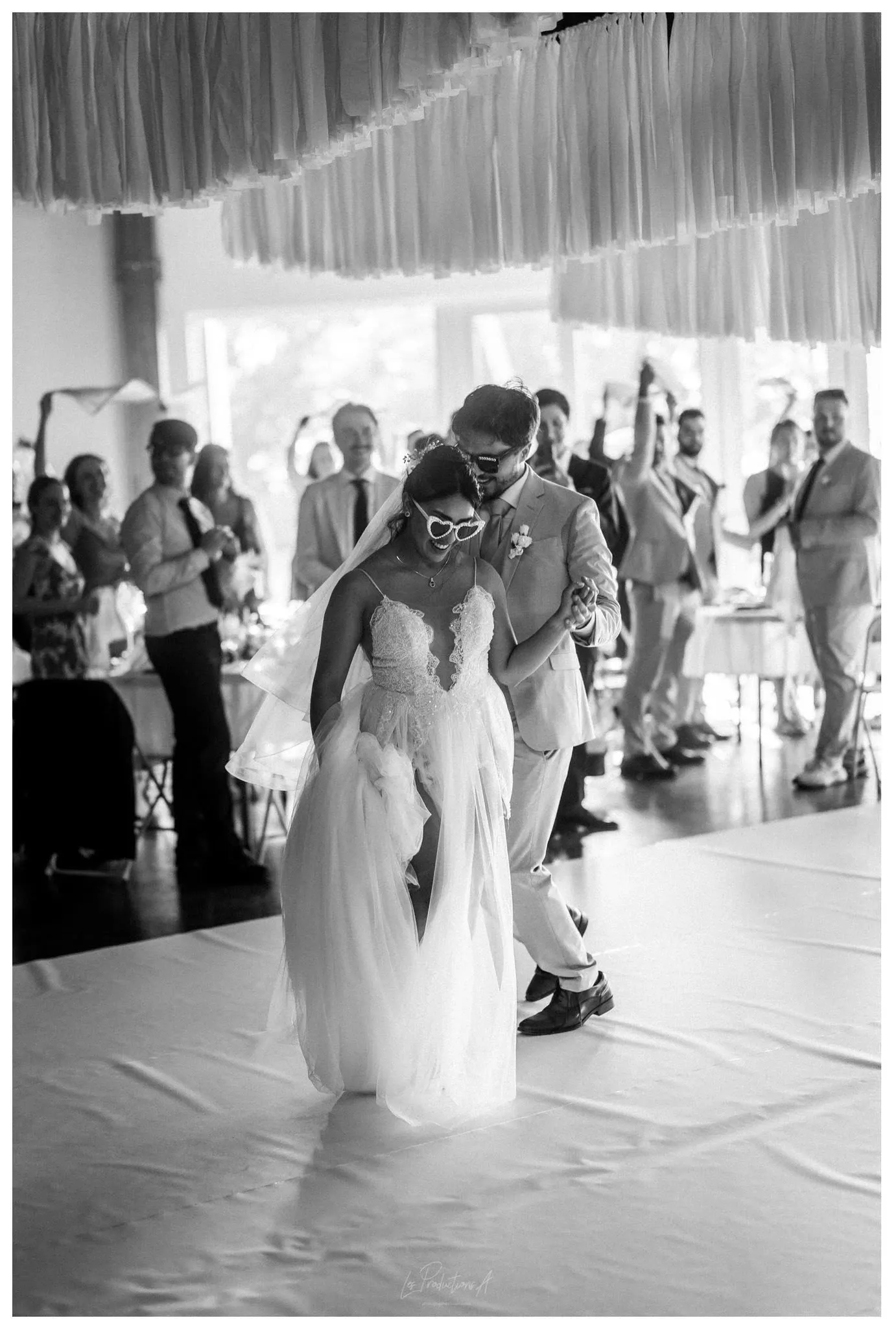 This black and white image, photographed by Les Productions A, captures a joyful moment from a wedding celebration, showcasing their expertise in wedding photography in Canada. The bride and groom, elegantly dressed, are dancing closely together. The bride is wearing a flowing gown and sunglasses, adding a playful touch to the moment, while the groom, in a stylish suit, appears delighted. In the background, wedding guests are gathered, some clapping and celebrating the couple's joyful occasion. This lively scene is set in a venue with soft, fabric decorations hanging from the ceiling, enhancing the festive atmosphere.