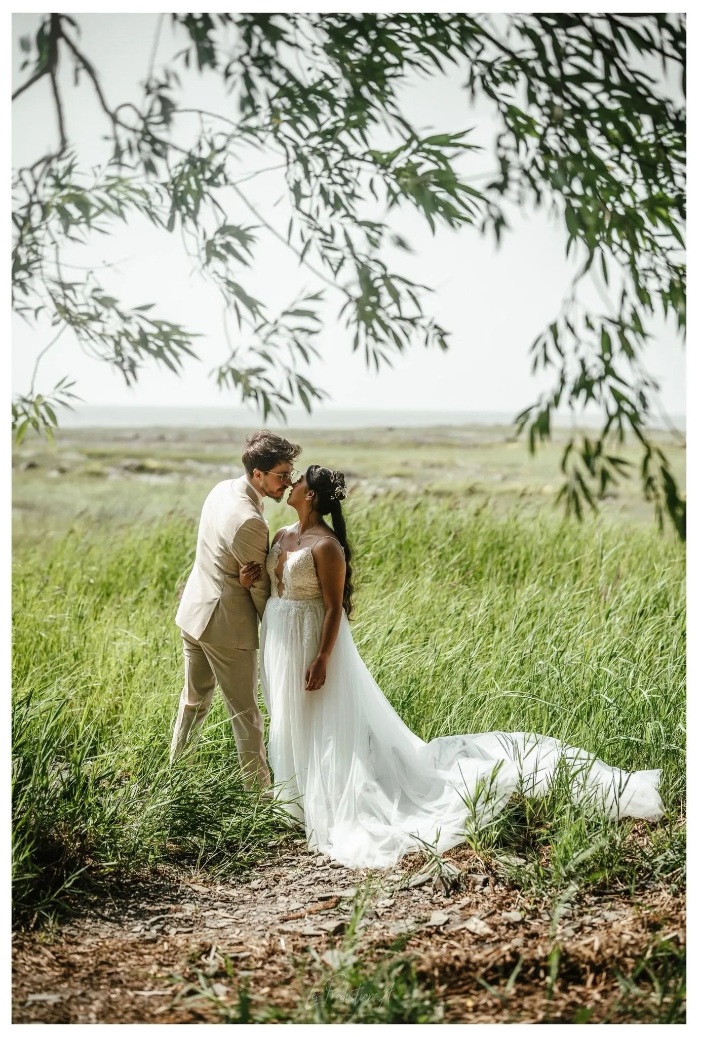 A bride and groom share a romantic moment in a lush, green meadow. The groom, wearing a beige suit, gently holds the bride, who is dressed in an elegant white wedding gown. They stand amidst tall grasses, with tree branches delicately framing the top of the image. The scene is serene and intimate, capturing the joy of their special day. This image is a display of wedding photography by Les Productions A, highlighting their talent in capturing natural and beautiful moments, suitable for the Mariage Elegant vendor listing in Canada.