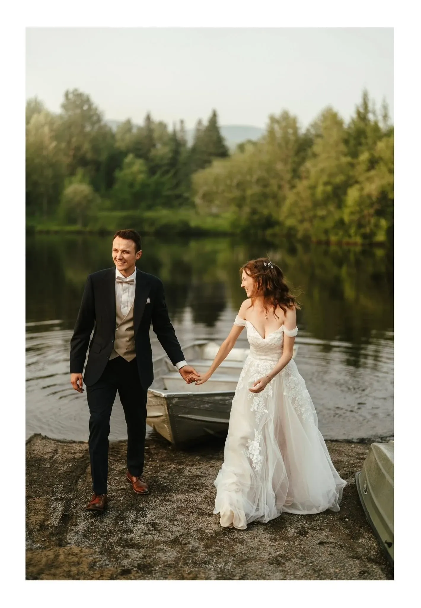 A newlywed couple is captured walking hand in hand by a serene lakeside, surrounded by lush greenery. The groom is wearing a classic dark suit with a bow tie, while the bride is in a flowing, off-the-shoulder white wedding gown adorned with lace details. The atmosphere is romantic and tranquil, with gentle ripples on the water reflecting the soft light of the setting sun. This image is part of the wedding photography portfolio by Les Productions A, showcasing natural and intimate moments for Mariage Elegant in Canada.