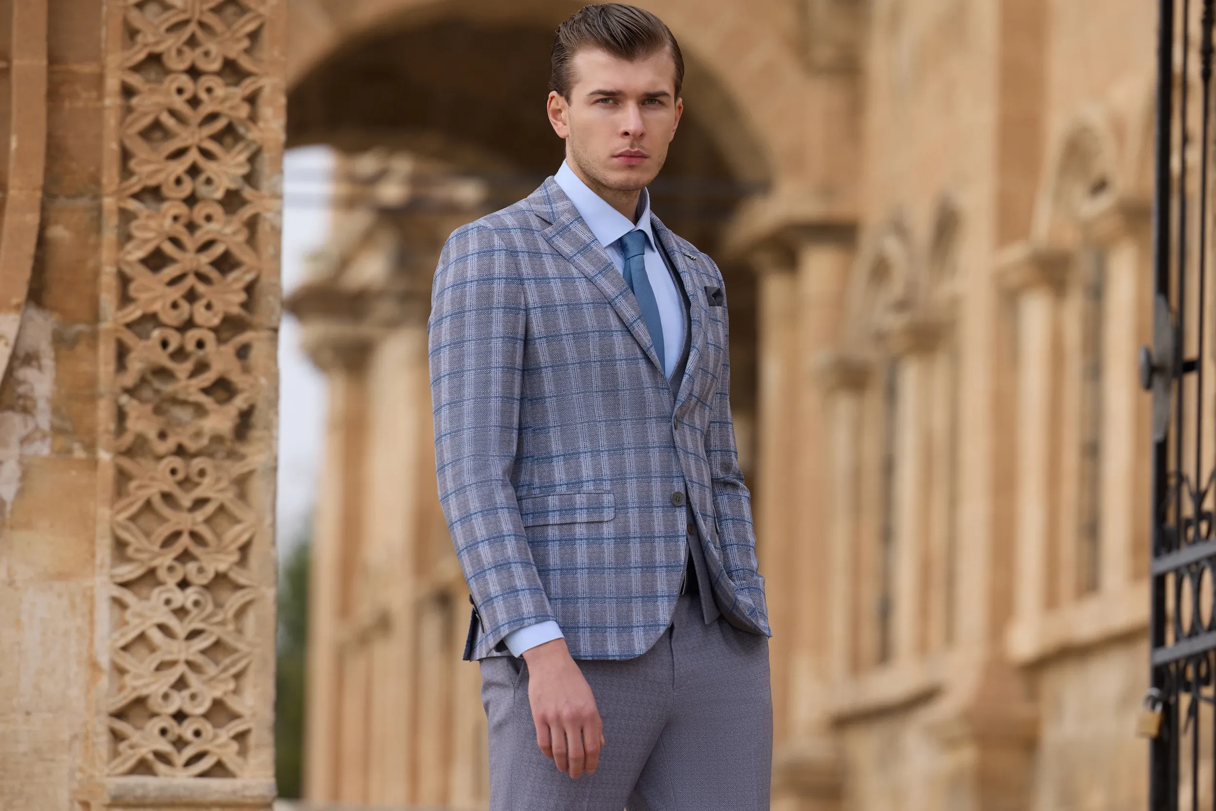 A well-dressed man stands confidently in an elegantly designed outdoor setting, showcasing a stylish wedding tuxedo by Baggi. The outfit features a light blue plaid jacket paired with matching light gray trousers, complemented by a crisp white shirt and a coordinated blue tie. The architectural backdrop of stone arches and intricate patterns adds a touch of sophistication, perfectly aligning with the upscale wedding theme featured by Mariage Elegant in Canada.