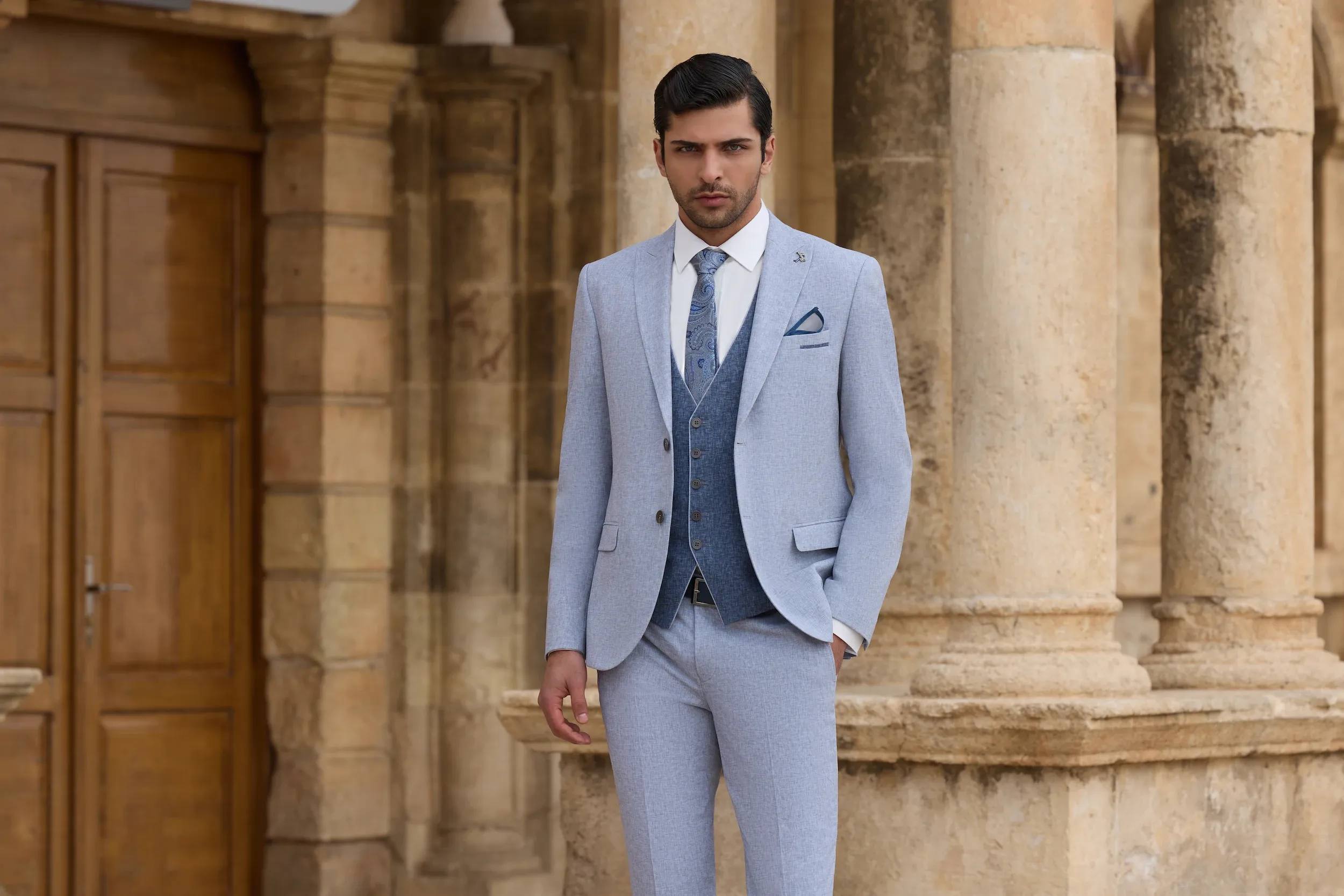 A groom stands confidently in front of an elegant stone building, showcasing a stylish wedding tuxedo by Baggi. The tuxedo features a light grey jacket and matching trousers, complemented by a deep blue vest and a patterned tie, ideal for a sophisticated wedding look. This attire, part of the Wedding Tuxedo category, is perfect for those seeking a modern yet classic style for their special day. The tranquil outdoor setting emphasizes the timeless elegance of the ensemble, fitting for a memorable wedding event in Canada.