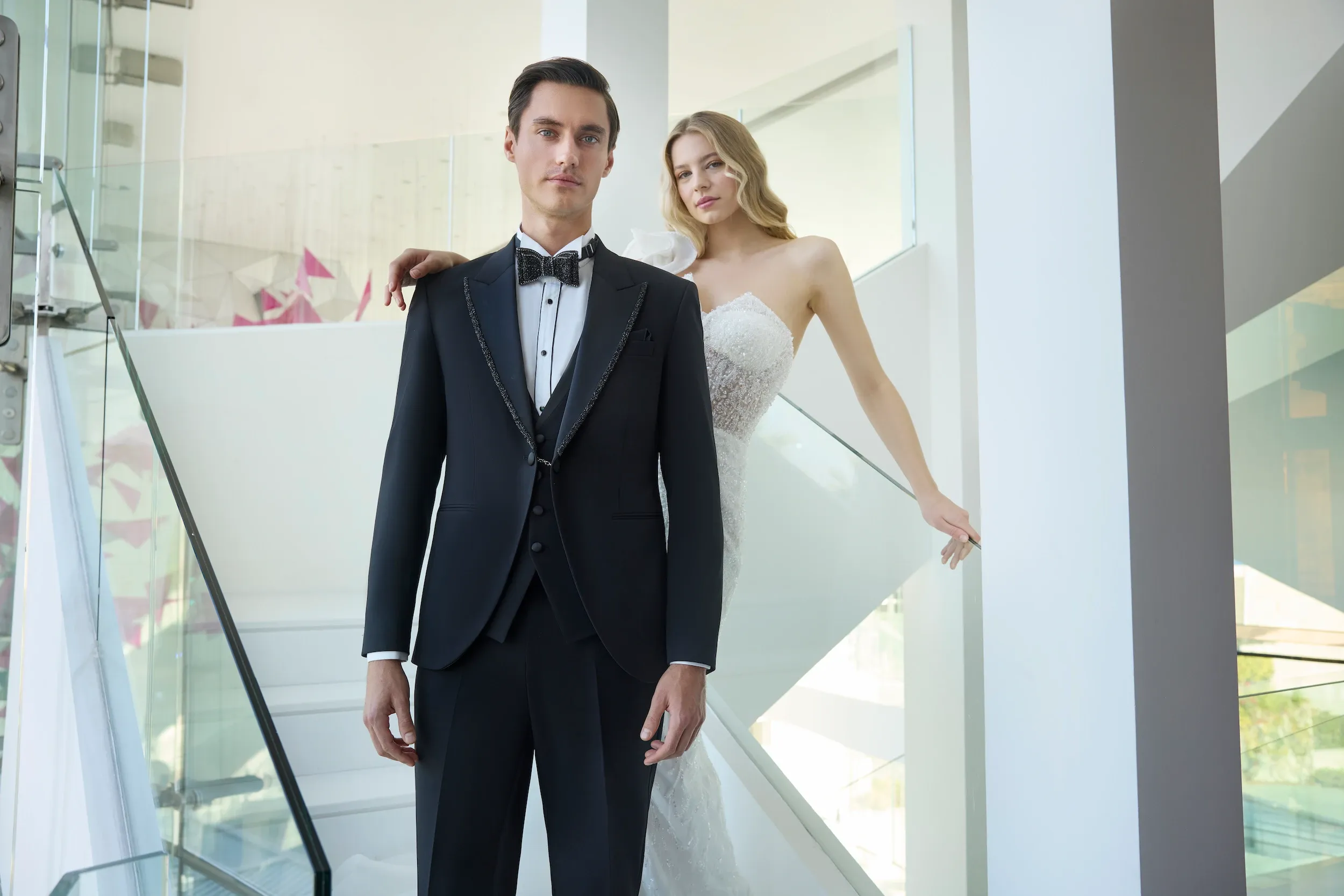 A groom wearing an elegant, tailored black tuxedo from Baggi's wedding tuxedo collection stands confidently in a modern venue, which is part of the Mariage Elegant, Canada setting. The tuxedo features a classic bow tie and intricate detailing. Beside him, a bride in a stunning white lace gown gently holds his shoulder, enhancing the formal and sophisticated atmosphere. They are posed in a bright, contemporary space with clean lines and glass accents, ideal for a stylish wedding ceremony.