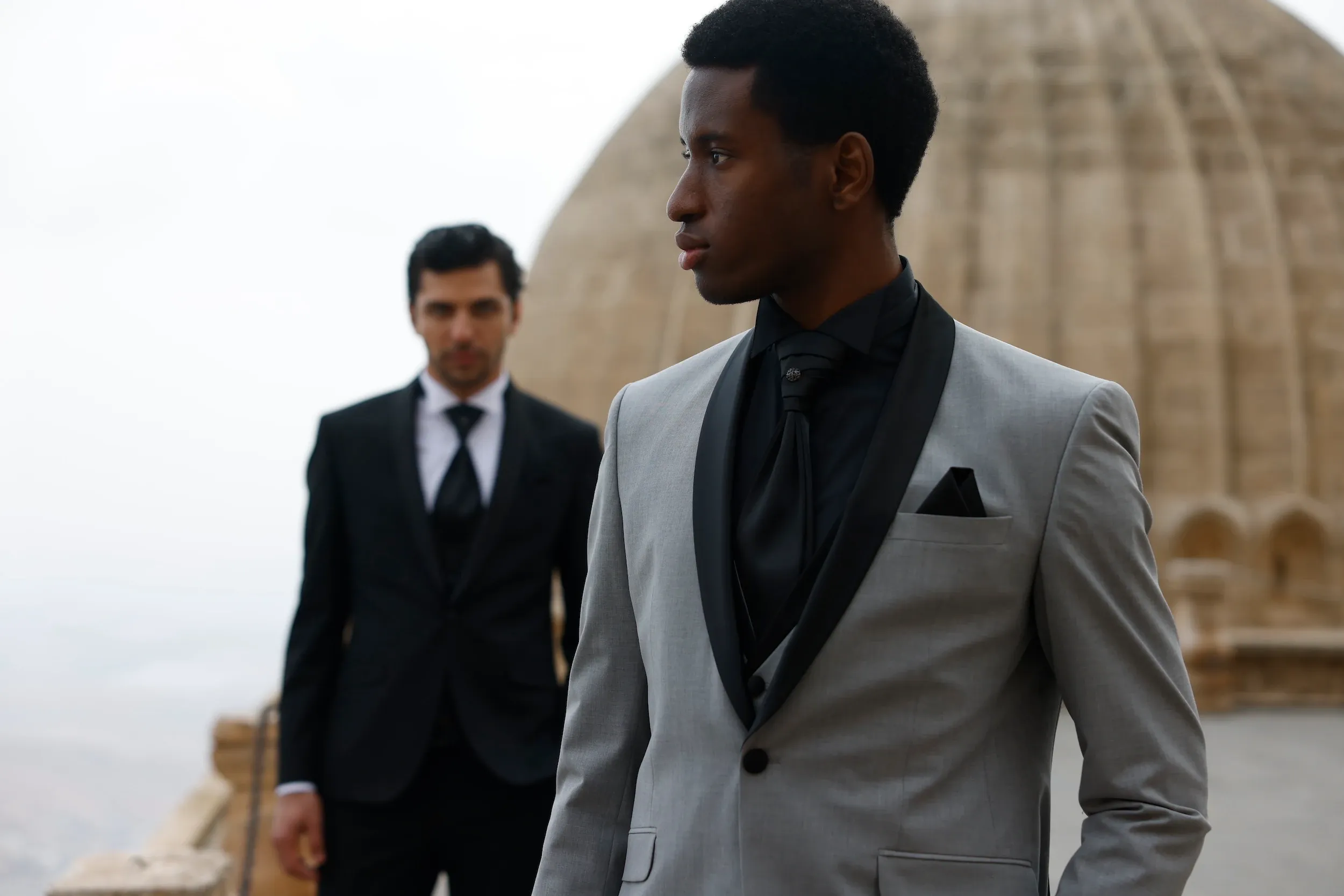 Two men are elegantly dressed in wedding attire, highlighting options from Baggi in the Wedding Tuxedo category. The man in the foreground wears a stylish light gray tuxedo with a contrasting black lapel, paired with a black dress shirt and a matching black tie. His pocket square adds a sophisticated touch. In the background, another man is dressed in a classic black tuxedo with a white dress shirt and a black tie. They are positioned outdoors against a backdrop of what appears to be an ancient stone dome, adding an element of timeless elegance suitable for a wedding setting.