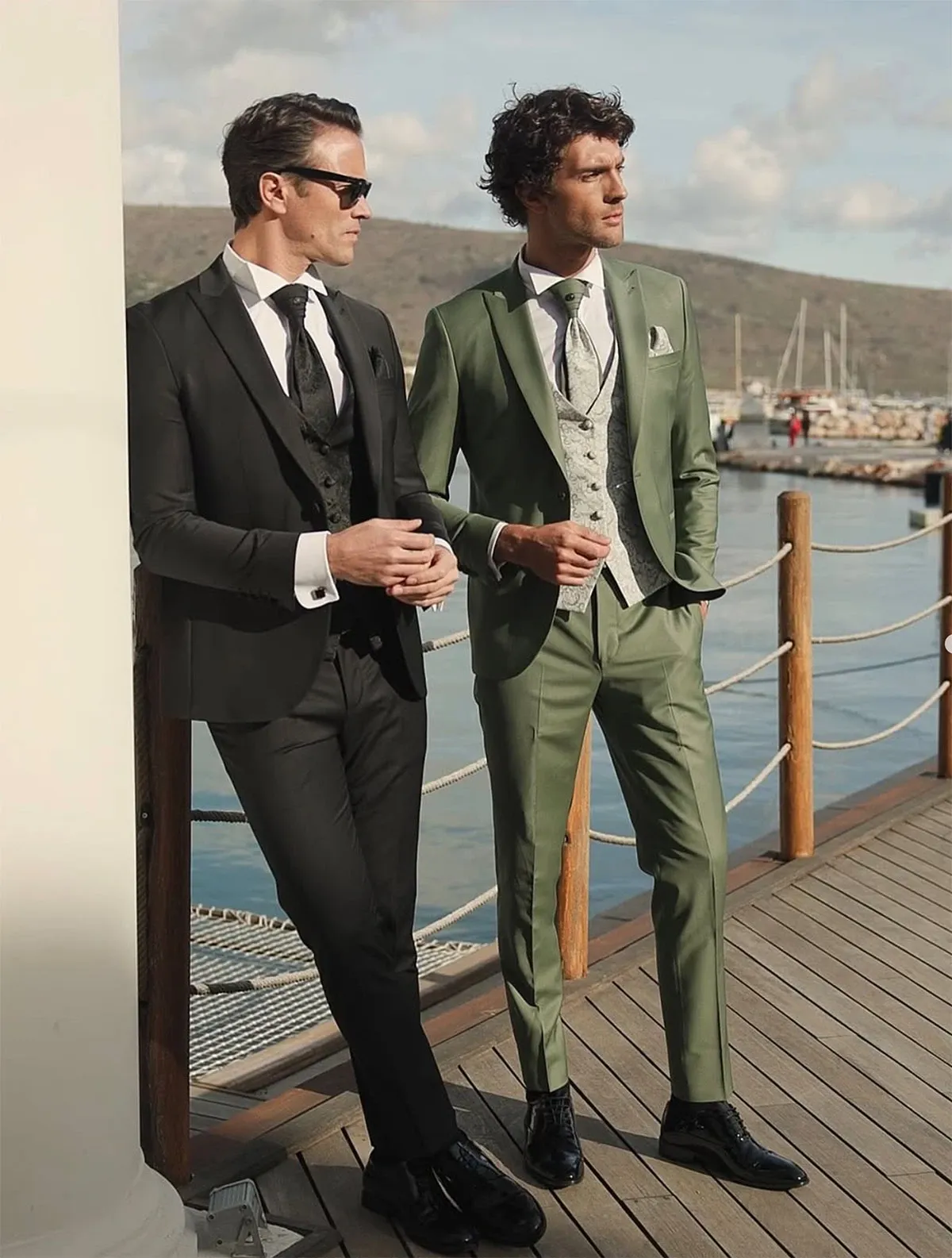 Two elegantly dressed men stand by a waterfront, showcasing wedding tuxedos from Baggi. The man on the left is wearing a classic black tuxedo with a shiny satin lapel, a crisp white shirt, a black waistcoat, and a black tie, completed with polished black shoes and stylish sunglasses. The man on the right sports a modern, light olive green tuxedo with a matching waistcoat, a white shirt, and a black and white patterned tie, paired with shiny black shoes. Both men are positioned casually against a rope railing, with a scenic marina and hills in the background, emphasizing a sophisticated wedding attire display from the vendor Baggi, in the category of Wedding Tuxedo.