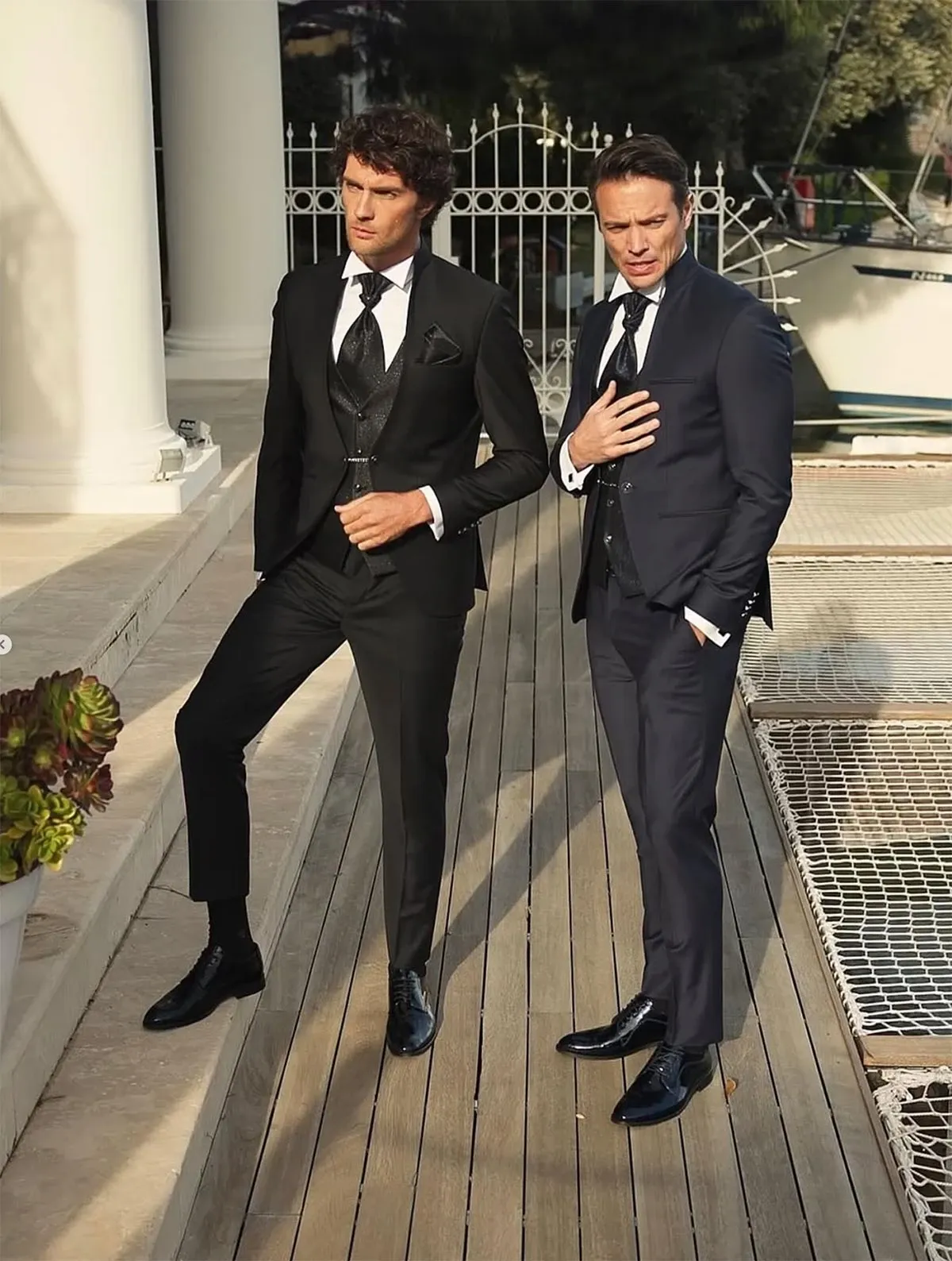 Two elegantly dressed men are showcasing wedding tuxedos by Baggi, a featured vendor in the Wedding Tuxedo category on Mariage Elegant, Canada. The image displays one man in a classic black tuxedo, complete with a matching vest, crisp white shirt, and black tie, while the other wears a sophisticated navy blue tuxedo with similar accompaniments. Both tuxedos are complemented by polished black dress shoes. The setting appears to be an upscale outdoor venue with a wooden deck, white columns, and wrought iron fencing, adding a touch of luxury to the wedding attire presentation.