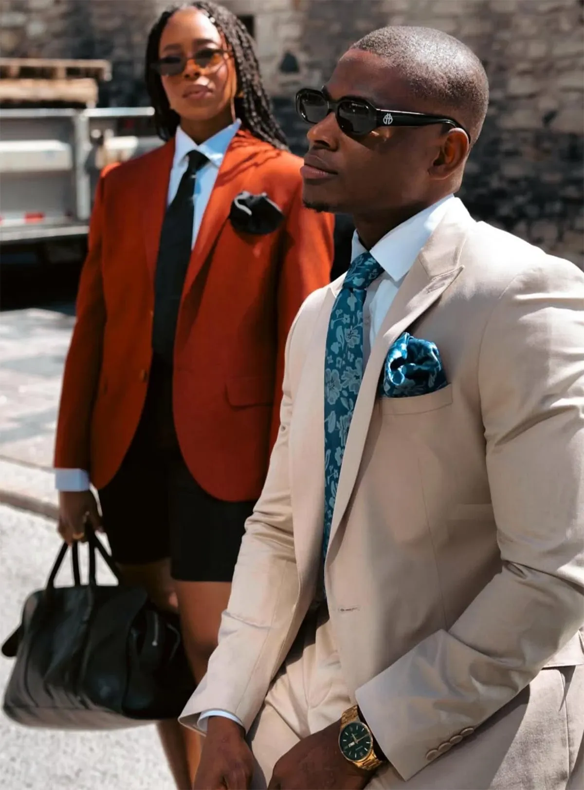 In this image, two individuals are elegantly dressed in sophisticated attire suitable for a wedding. The focus is on the person in the foreground, wearing a sleek, light-colored tuxedo with a teal floral tie and a matching pocket square that adds a touch of style. Completing the look is a gold watch that enhances the overall elegance. Behind them, another individual is dressed in a stylish red blazer paired with a white shirt, black tie, and black shorts, holding a black leather bag. The setting appears to be an outdoor urban environment with a stone wall in the background. This image is part of the wedding tuxedo category for the vendor Baggi, featured on the Mariage Elegant platform in Canada, showcasing fashionable options for wedding attire.