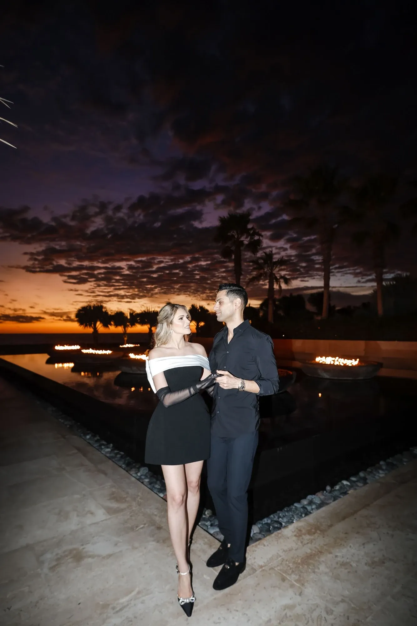 A couple stands elegantly next to a serene water feature at sunset, captured by Tiara Studio, showcasing their expertise in wedding photography. The sky above is painted with dramatic hues of purple and orange, reflecting the romantic ambiance perfect for wedding portraits. The woman wears a stylish black and white dress, while the man is in a sophisticated dark outfit. This image highlights Tiara Studio's ability to blend natural beauty with captivating moments, ideal for couples looking to capture their special day in a picturesque setting.