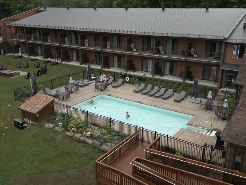 Cette image montre une grande salle de réception à deux étages avec un toit en métal et des balcons individuels. Devant le bâtiment, il y a une piscine rectangulaire entourée de chaises longues et de parasols. Quelques personnes se détendent autour de la piscine, créant une atmosphère détendue et accueillante. Un jardin bien entretenu entoure la piscine, ajoutant une touche de verdure et d'élégance à l'ensemble. Cette salle de réception est idéale pour organiser des événements de mariage en plein air, combinant l'hébergement et des installations de loisirs pour les invités.