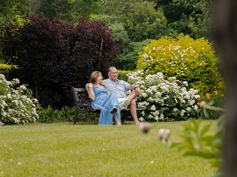 Un couple est assis sur un banc en fer forgé dans un vaste jardin verdoyant, entouré de fleurs blanches et de buissons colorés. La femme porte une robe bleue élégante, tandis que l'homme est en tenue décontractée avec un short beige et une chemise claire. Le jardin, idéal pour des événements en extérieur, offre un cadre paisible et romantique, parfait pour des célébrations de mariage en plein air. La verdure luxuriante et les fleurs ajoutent une touche naturelle et sereine à l'atmosphère.