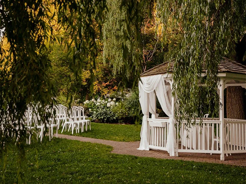 Cette image montre un charmant espace de mariage en plein air, situé dans un cadre verdoyant. Au centre se trouve une gloriette en bois blanc décorée de rideaux légers et vaporeux, ajoutant une touche romantique et élégante. À gauche, des rangées de chaises blanches sont disposées pour accueillir les invités, indiquant la préparation pour une cérémonie de mariage. L'environnement est entouré de feuillage luxuriant et de fleurs blanches florissantes, évoquant une atmosphère paisible et naturelle. Cet espace est idéal pour un mariage intime et élégant, offrant une belle harmonie avec la nature environnante.