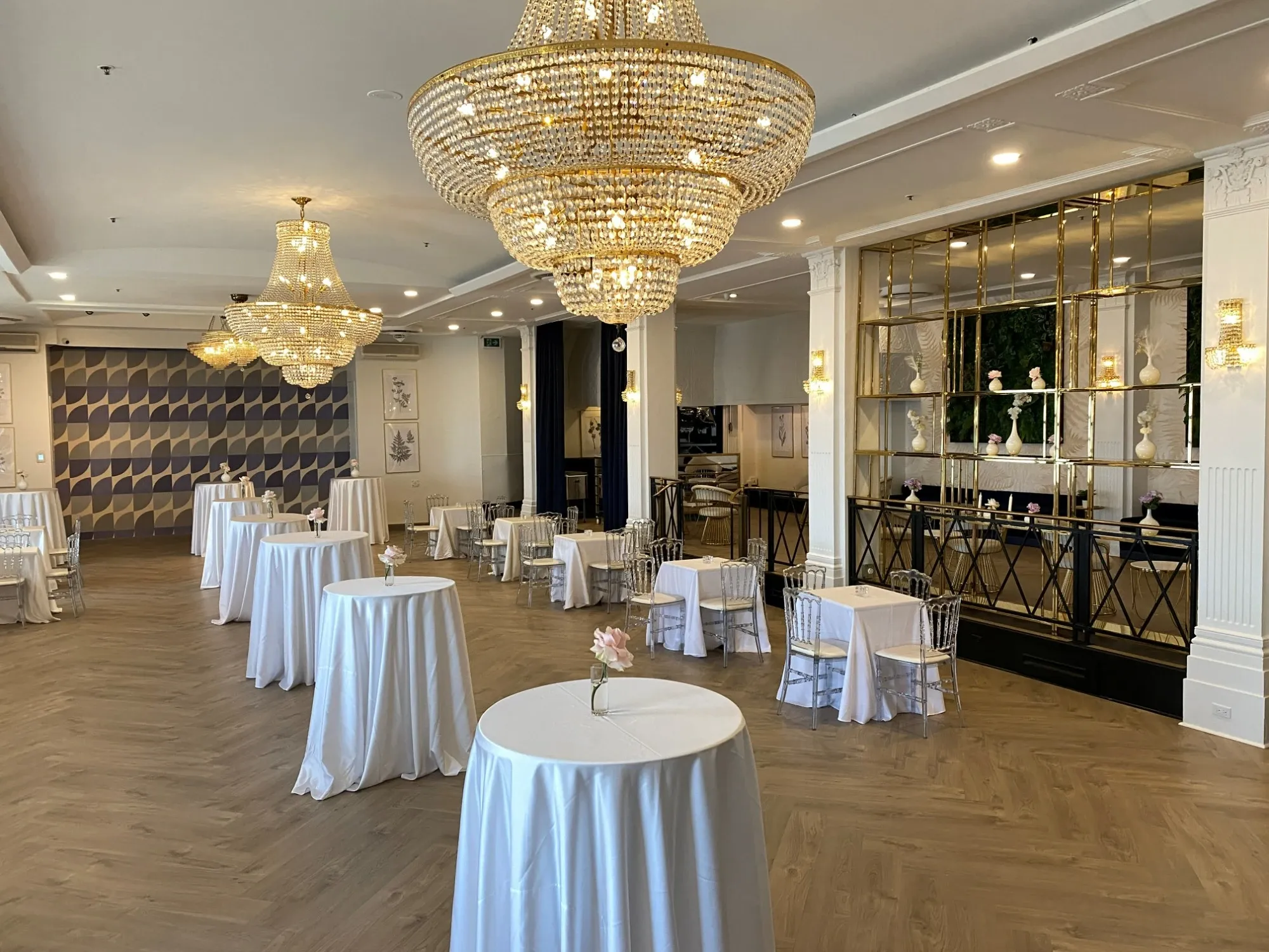 The image shows an elegant wedding venue provided by The PMG Group, featured under the wedding venues category on Mariage Elegant, Canada. The spacious room is adorned with luxurious chandeliers that cast a warm glow over the setting. Round tables covered with crisp white tablecloths are evenly spaced throughout the venue, each topped with a delicate floral centerpiece. The floor is a polished wood, adding a touch of sophistication. In the background, a stylish, patterned wall and a mirrored bar area enhance the venue's luxurious ambiance, ideal for hosting exquisite weddings.