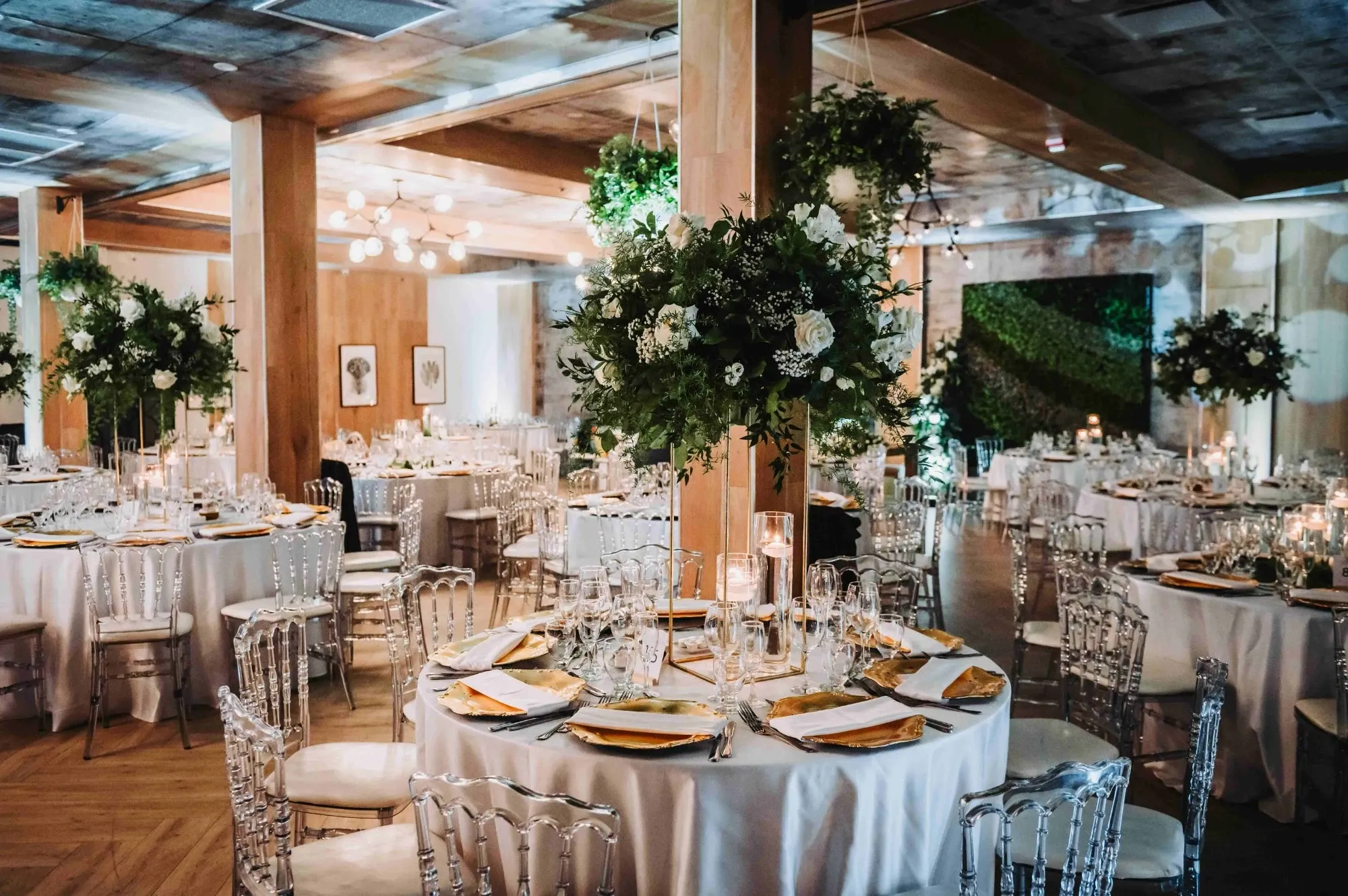The image showcases an elegant wedding venue by The PMG Group, categorized under Wedding Venues on Mariage Elegant, Canada. The venue features beautifully set round tables adorned with white tablecloths and gold-accented tableware. Lush green and white floral arrangements serve as centerpieces, complementing the modern, transparent chairs arranged around each table. Soft, ambient lighting creates a warm and inviting atmosphere, enhanced by the greenery wall in the background. This setting embodies sophistication and style, ideal for a memorable wedding celebration.