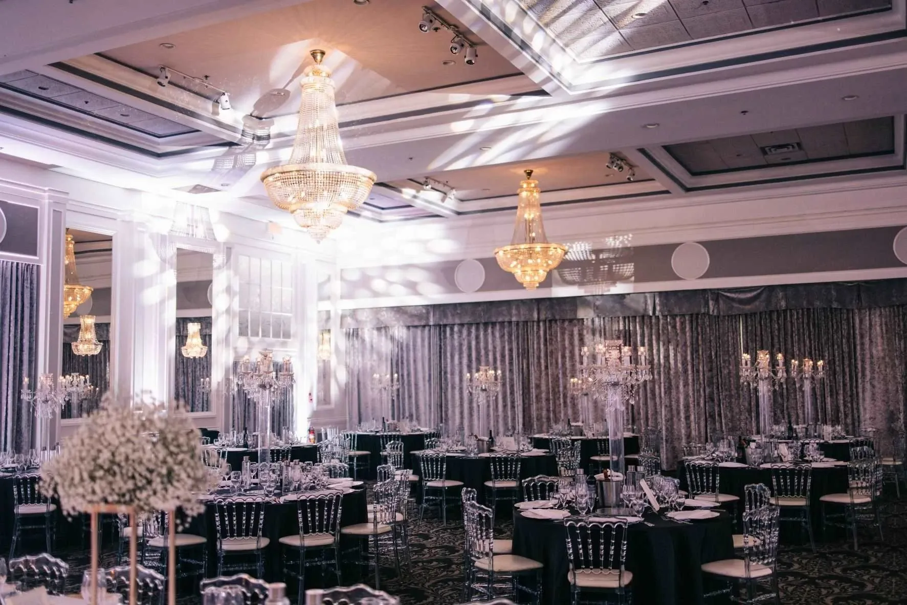 The image shows a luxurious wedding venue by The PMG Group, categorized under Wedding Venues, at Mariage Elegant in Canada. The room is elegantly decorated with crystal chandeliers hanging from a high, intricately designed ceiling. The walls are adorned with soft, shimmering drapes, and the floor is covered with a patterned dark carpet. Round tables are arranged neatly, each set with transparent chairs and tall, sophisticated floral centerpieces, creating a refined and romantic ambiance suitable for a wedding reception.