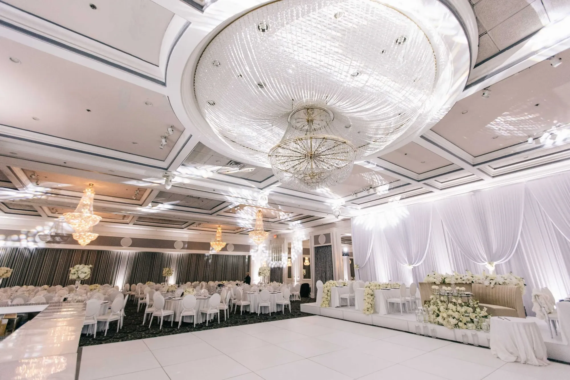 The image showcases an elegant wedding venue by The PMG Group, categorized under Wedding Venues, featured on Mariage Elegant, Canada. The luxurious hall is adorned with grand, sparkling chandeliers that illuminate the space with a warm glow. Round tables draped in pristine white tablecloths are neatly arranged, with chairs surrounding them, ready for guests. The venue is decorated with cascading white drapes and floral arrangements, highlighting the romantic ambiance suitable for weddings. A large, polished dance floor occupies the central space, inviting guests for celebration and dancing.