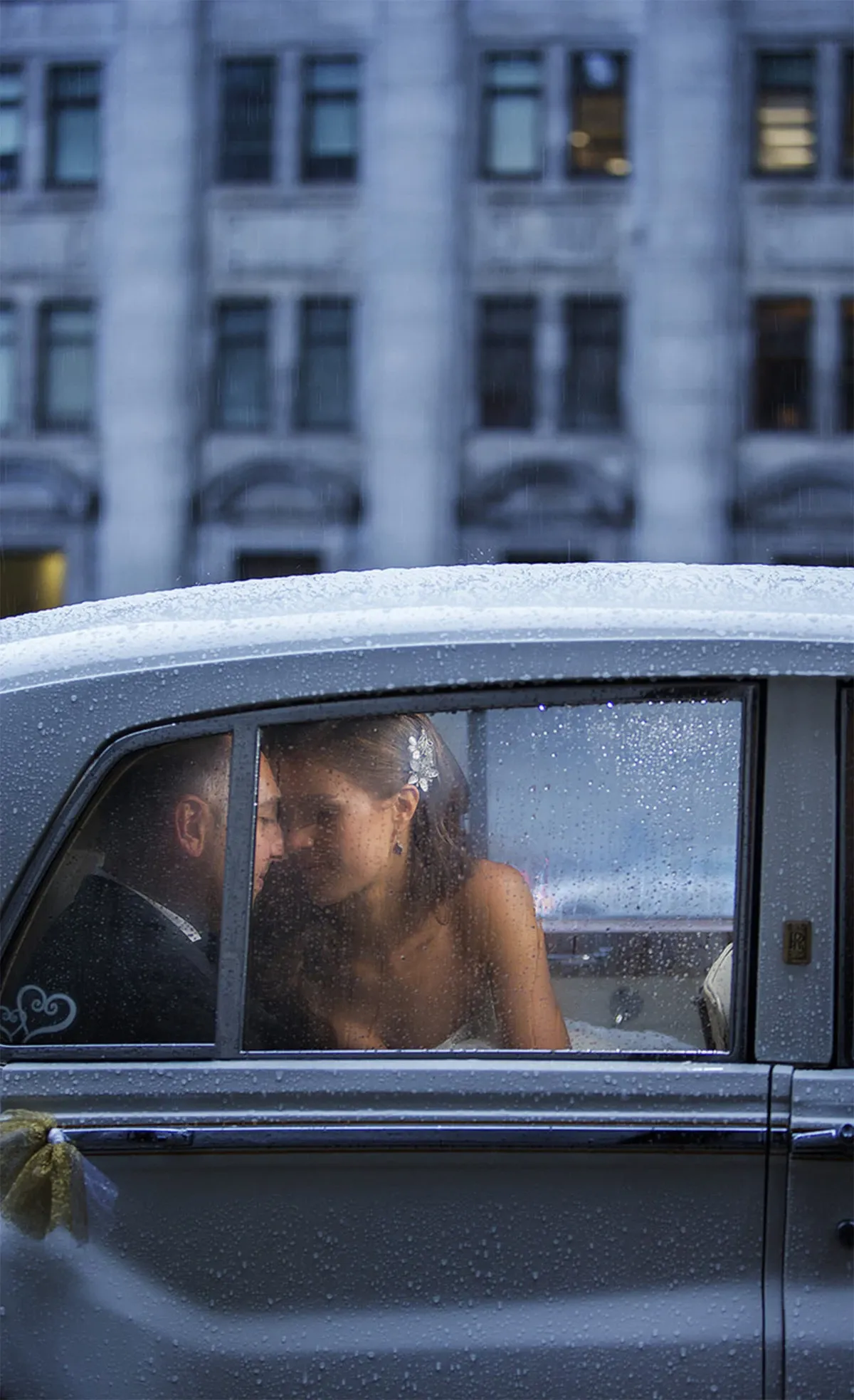 A bride and groom share an intimate moment inside a classic car with raindrops on the window, adding a romantic touch to the scene. The bride is wearing an elegant strapless wedding dress and a delicate hair accessory. The cityscape outside displays the facade of a historic building, suggesting an urban setting. This image captures a candid moment between the couple on their special day. Inhouse Studios Photography, specializing in wedding photography, beautifully encapsulates this tender moment for Mariage Elegant's vendor listing in Canada.