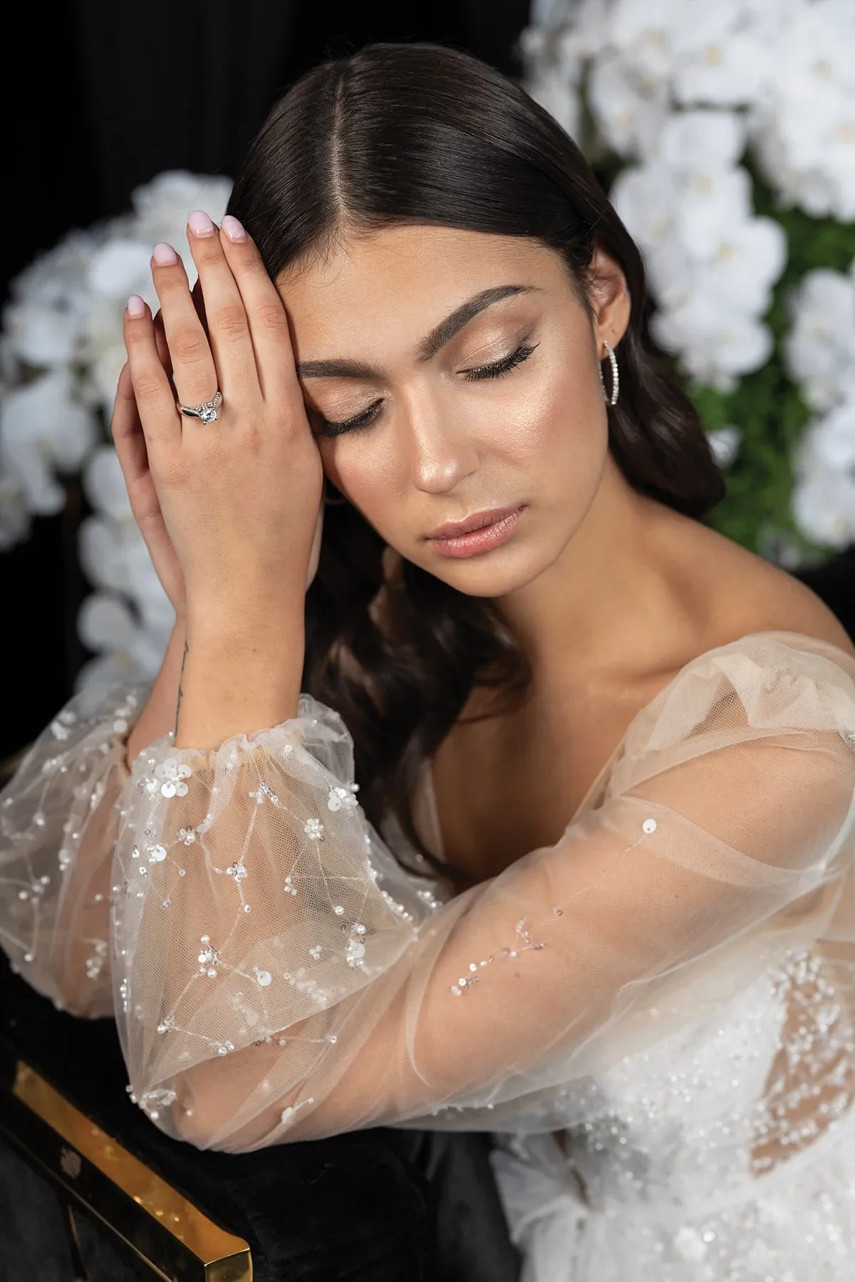 Une mariée élégante aux cheveux bruns et au maquillage subtil ferme les yeux doucement. Elle porte une robe de mariée avec des manches en tulle ornées de délicates perles. Sa main gauche repose près de son visage, mettant en valeur une bague de fiançailles scintillante. En arrière-plan, de magnifiques fleurs blanches ajoutent une touche de romantisme. Cette image est idéale pour illustrer la catégorie des bijoux de mariage, soulignant la beauté et le raffinement des accessoires pour le grand jour.