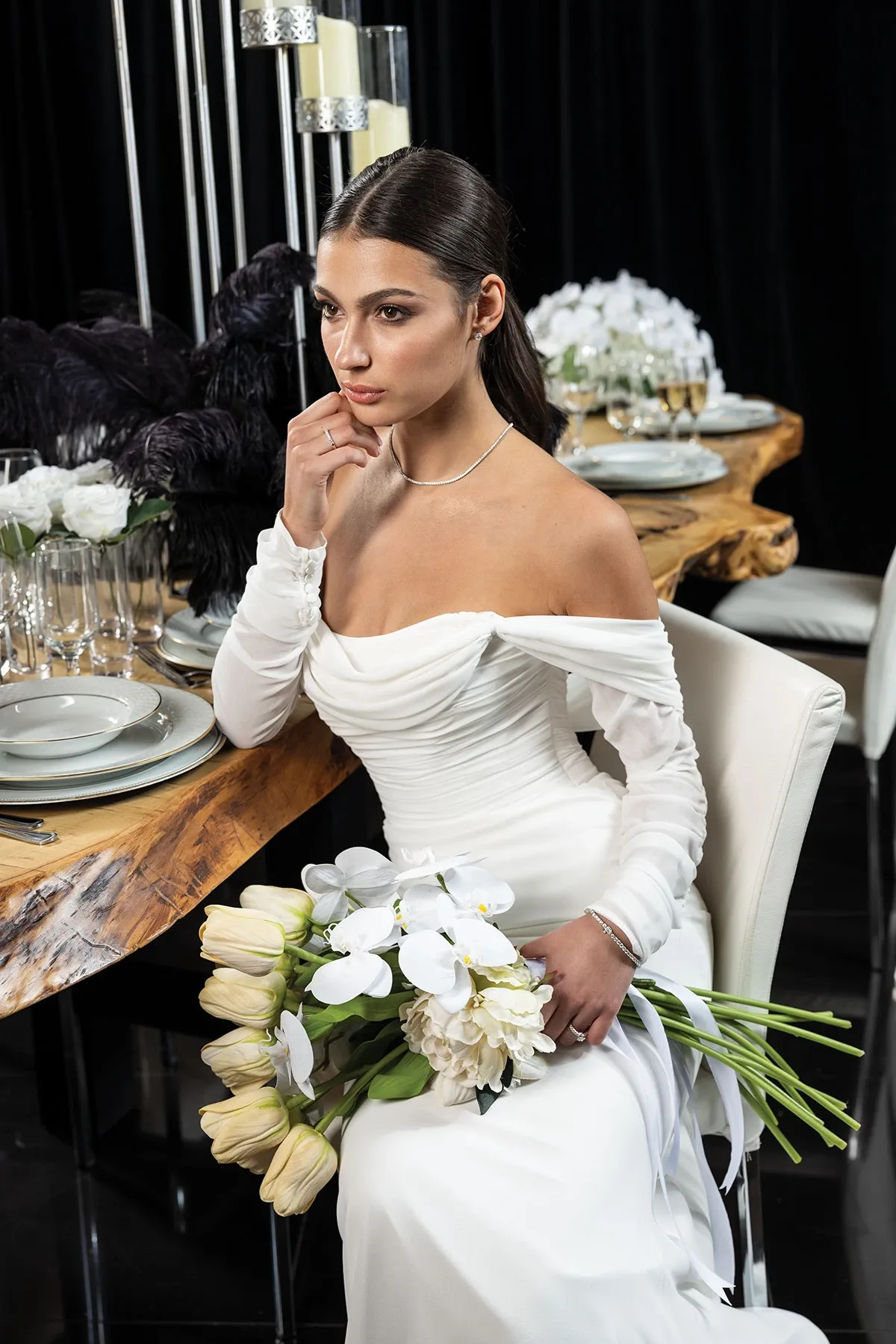 Une femme en robe de mariée blanche élégante est assise à une table soigneusement décorée pour un mariage. Elle porte un collier délicat et tient un bouquet de tulipes blanches et d'orchidées. La table est ornée de vaisselle raffinée et de compositions florales sophistiquées, créant une ambiance chic et romantique. Des éléments de décoration modernes, tels que des chandeliers en métal, ajoutent une touche contemporaine à l'ensemble. Cette image illustre magnifiquement les options de bijoux et de décorations florales pour un mariage luxueux.