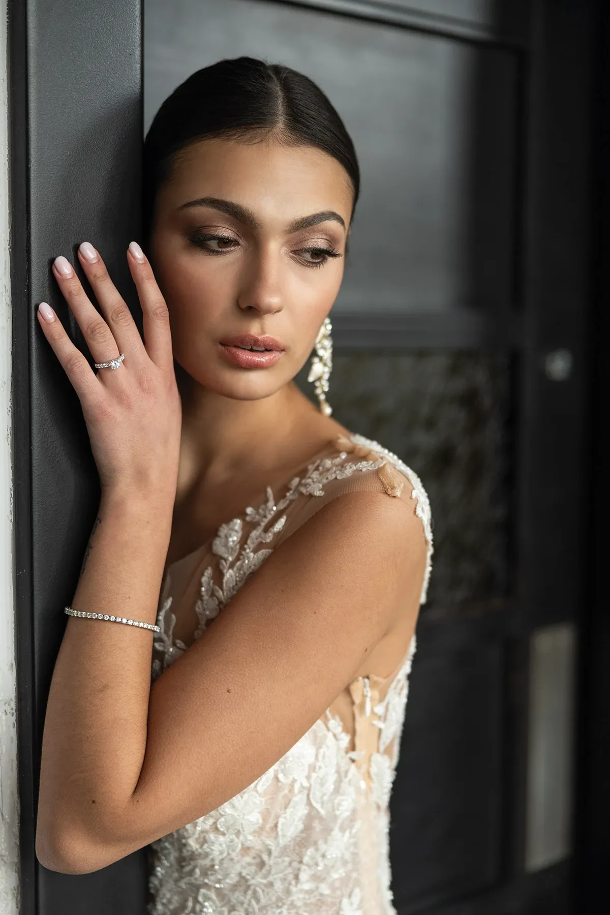 Une mariée élégante porte une robe de mariage délicatement ornée de dentelle avec des motifs floraux. Elle est appuyée contre un mur, regardant pensivement au loin. Ses cheveux sont soigneusement coiffés en un chignon bas sophistiqué. La mariée arbore un maquillage naturel mettant en valeur ses traits gracieux. Elle porte une bague de fiançailles étincelante en diamant à l'annulaire gauche et un bracelet fin en argent, complété par des boucles d'oreilles pendantes sophistiquées. Cette image incarne l'élégance et le raffinement des bijoux de mariage disponibles sur Mariage Elegant, Canada.