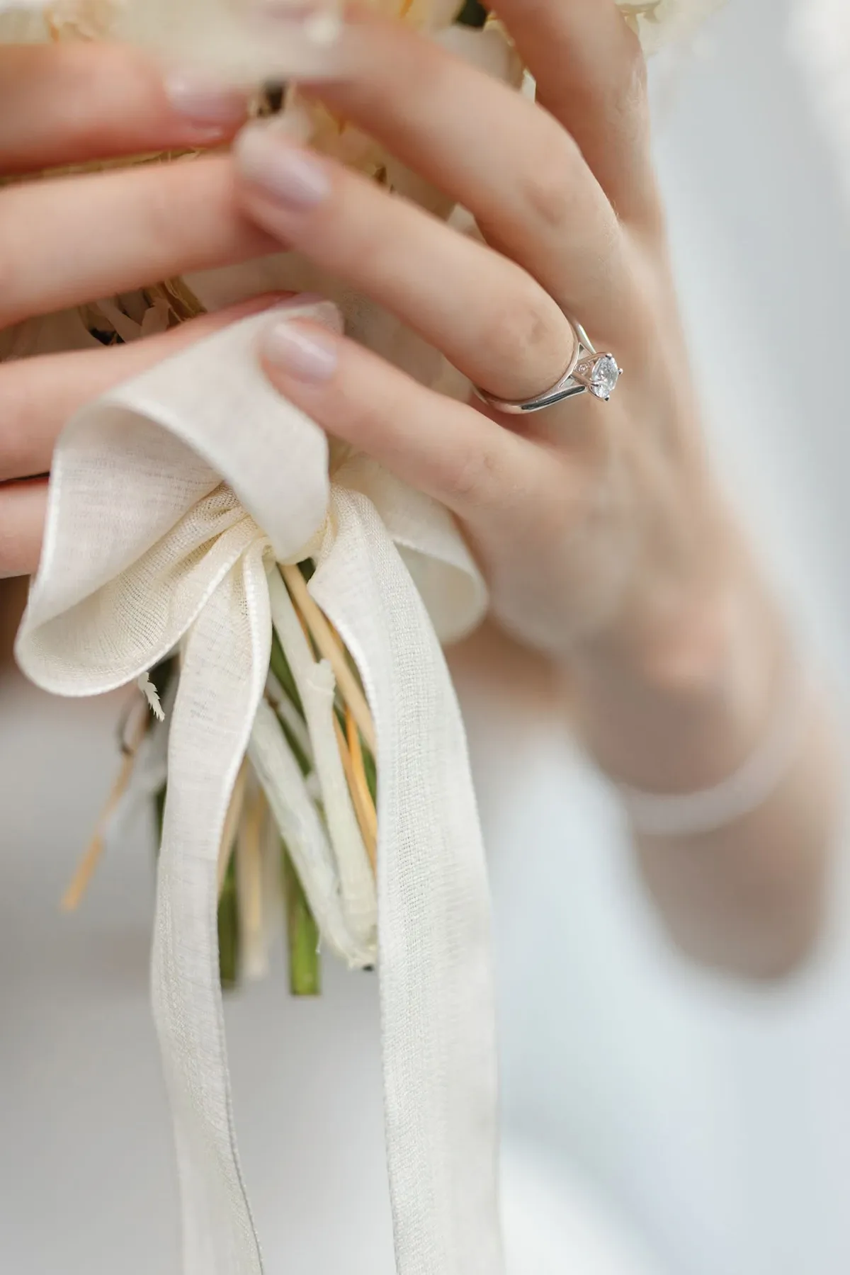 Une mariée tient délicatement un bouquet de fleurs blanches, accentué d'un ruban en tissu ivoire noué en boucle. Ses ongles sont manucurés dans une teinte rose pâle. À son doigt, elle porte une élégante bague de fiançailles en or blanc ornée d'un diamant scintillant, ajoutant une touche de raffinement et de glamour à cette scène de mariage.