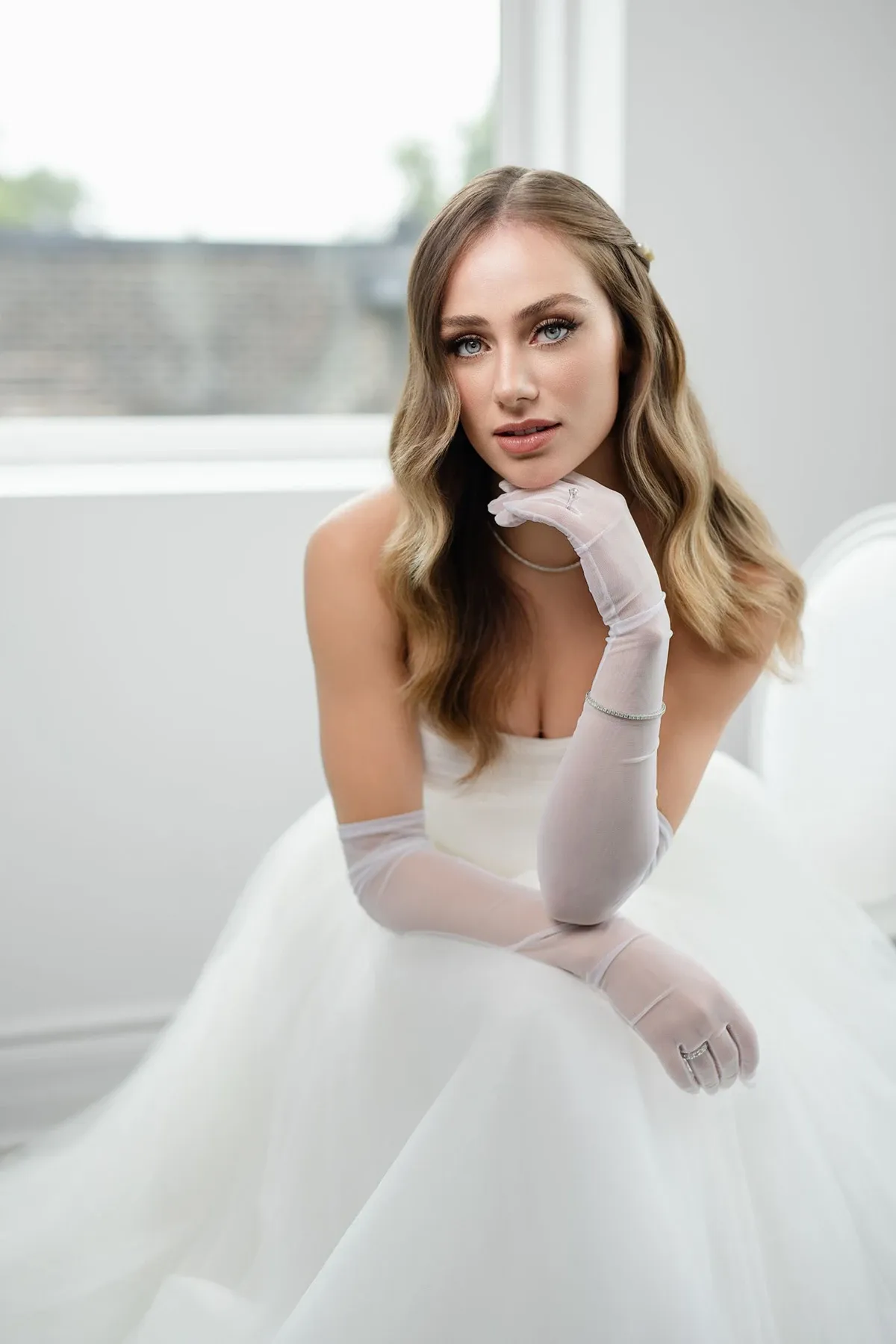 Une mariée élégante est assise près d'une fenêtre lumineuse, portant une robe de mariée blanche classique. Ses cheveux ondulés tombent délicatement sur ses épaules, et elle porte de longs gants transparents qui ajoutent une touche sophistiquée à son look nuptial. Son expression sereine et la lumière naturelle qui l'entoure créent une ambiance romantique et raffinée, idéale pour un mariage chic. Cette image est parfaite pour illustrer le choix de bijoux et d'accessoires élégants pour le jour du mariage sur Mariage Elegant, Canada.