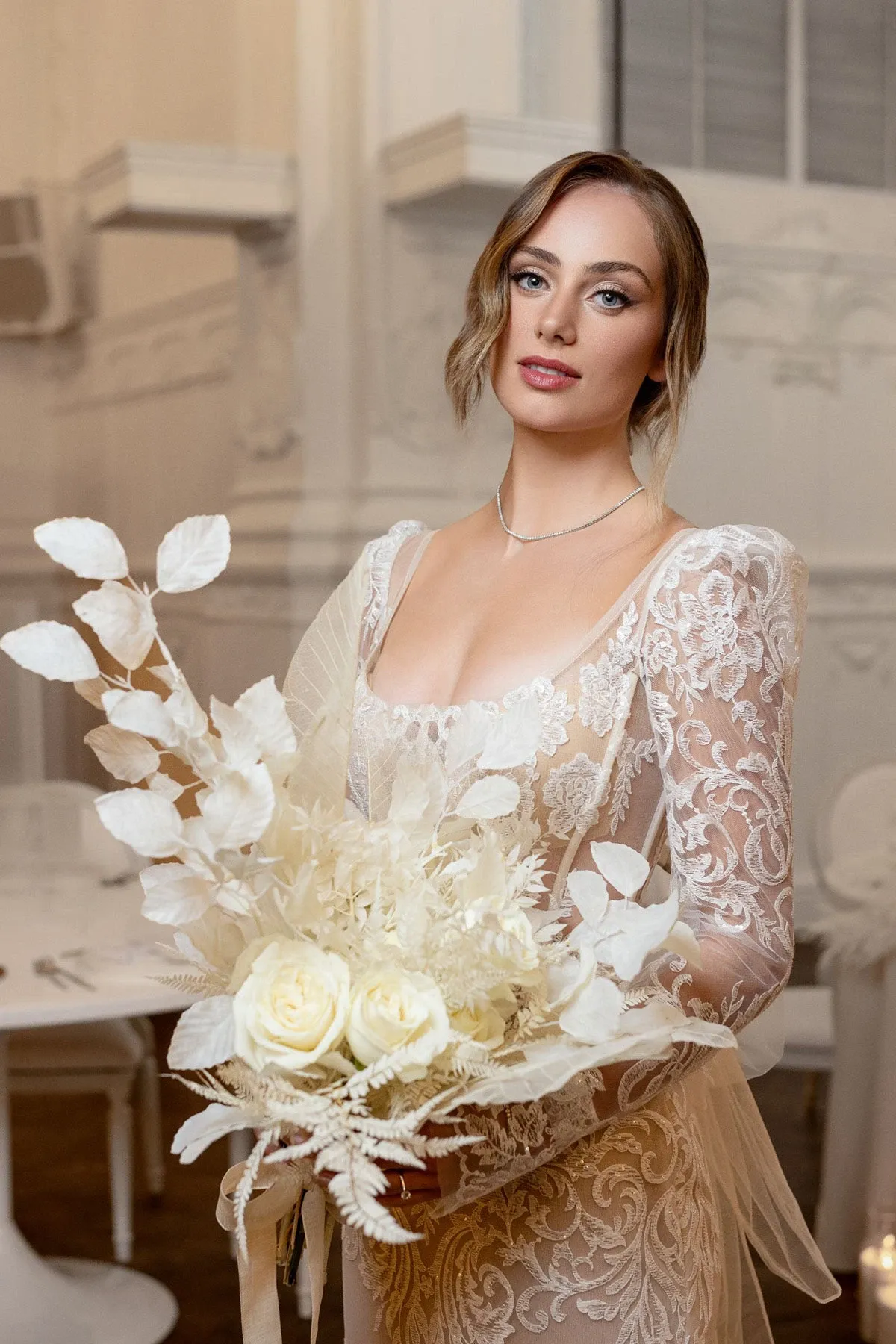 A bride stands elegantly in a tastefully decorated wedding venue, showcasing a delicate necklace that complements her attire. She wears an exquisite lace wedding dress with intricate floral patterns and long sleeves, exuding a timeless elegance. She holds a bridal bouquet composed of white roses, dried foliage, and delicate blooms, perfectly matching her gown's sophisticated design. The background features elegantly set tables, hinting at a formal and stylish wedding reception. This image is ideal for illustrating wedding jewelry options in a Canadian wedding planning context on the Mariage Elegant site.