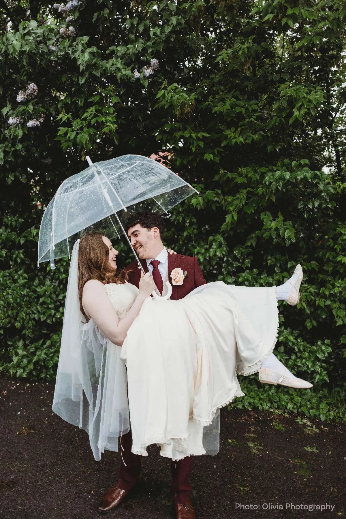 A groom in a burgundy suit playfully carries his bride, who is wearing an elegant white wedding gown with lace details, as they share a joyful moment. The bride holds a transparent umbrella overhead, hinting at a light rain. Both are smiling and appear delighted, reflecting the love and happiness of their special day. In the background, lush green foliage adds a natural, vibrant contrast to the scene, enhancing the romantic atmosphere. This image is exemplary of the joyful and intimate moments captured during wedding celebrations, showcasing the joyful union officiated by a skilled wedding officiant, crucial to creating memorable ceremonies.