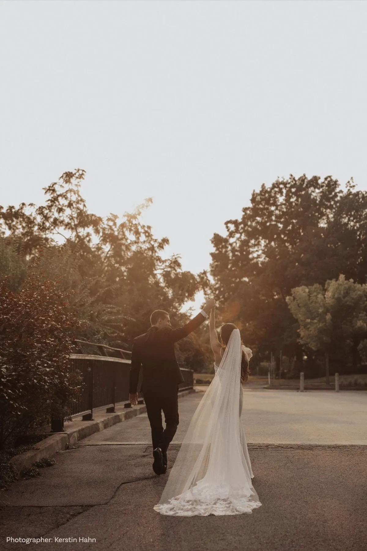 A newlywed couple is joyfully walking down a sunlit path, hand in hand, with the groom elegantly dressed in a black suit and the bride wearing a stunning white gown with a long, flowing veil. The evening sun casts a warm glow as they dance, creating a romantic atmosphere. Trees and foliage line the path, adding a natural touch to the serene setting. This image captures the couple's first moments as newlyweds after the ceremony, symbolizing the beginning of their journey together.