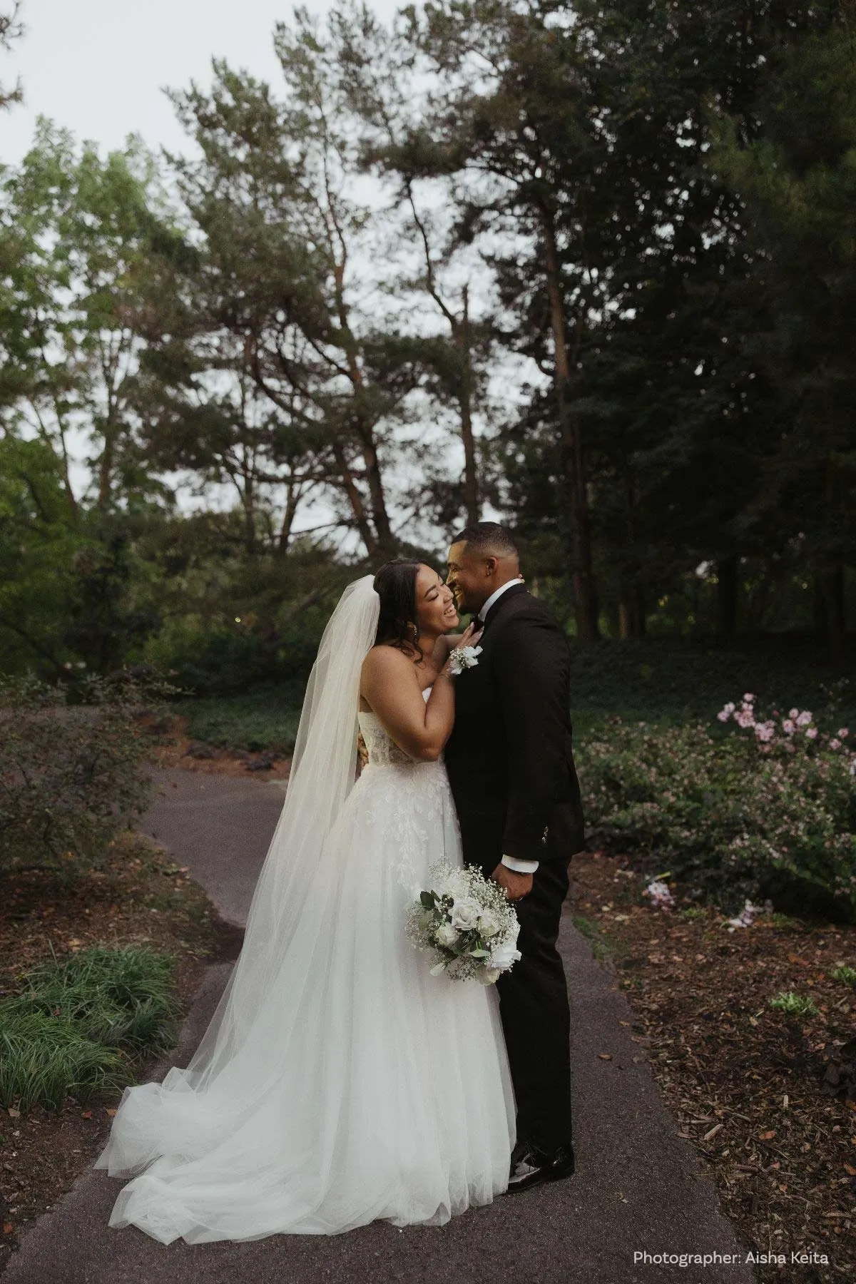 Un couple de mariés se tient tendrement sur un chemin bordé de verdure et de fleurs, un cadre idéal pour une cérémonie de mariage en extérieur. La mariée porte une robe blanche élégante avec un voile délicat, tenant un bouquet de fleurs blanches. Le marié, quant à lui, est vêtu d'un costume noir classique. Ils partagent un moment romantique sous les arbres majestueux, ajoutant une touche de magie à leur journée spéciale. Cette image illustre parfaitement l'amour et la joie d'un mariage célébré en pleine nature, soulignant l'importance de choisir le bon officiant pour ce moment unique.