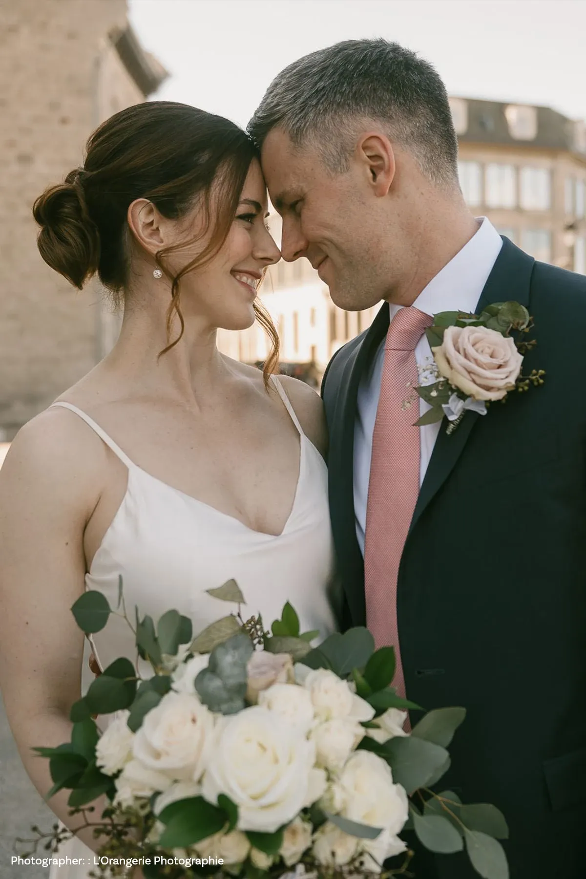 Un couple de mariés se tient proche l'un de l'autre, souriant et échangeant un regard complice devant un bâtiment en pierre sous un ciel clair. La mariée porte une robe blanche simple avec des bretelles et un chignon élégant, tandis que le marié est vêtu d'un costume bleu marine avec une cravate rose. Il a une boutonnière composée de roses assorties au bouquet de la mariée, qui est un arrangement élégant de roses blanches et de feuillage vert. Cette image émotive est idéale pour illustrer un célébrant de mariage sur Mariage Elegant, Canada.