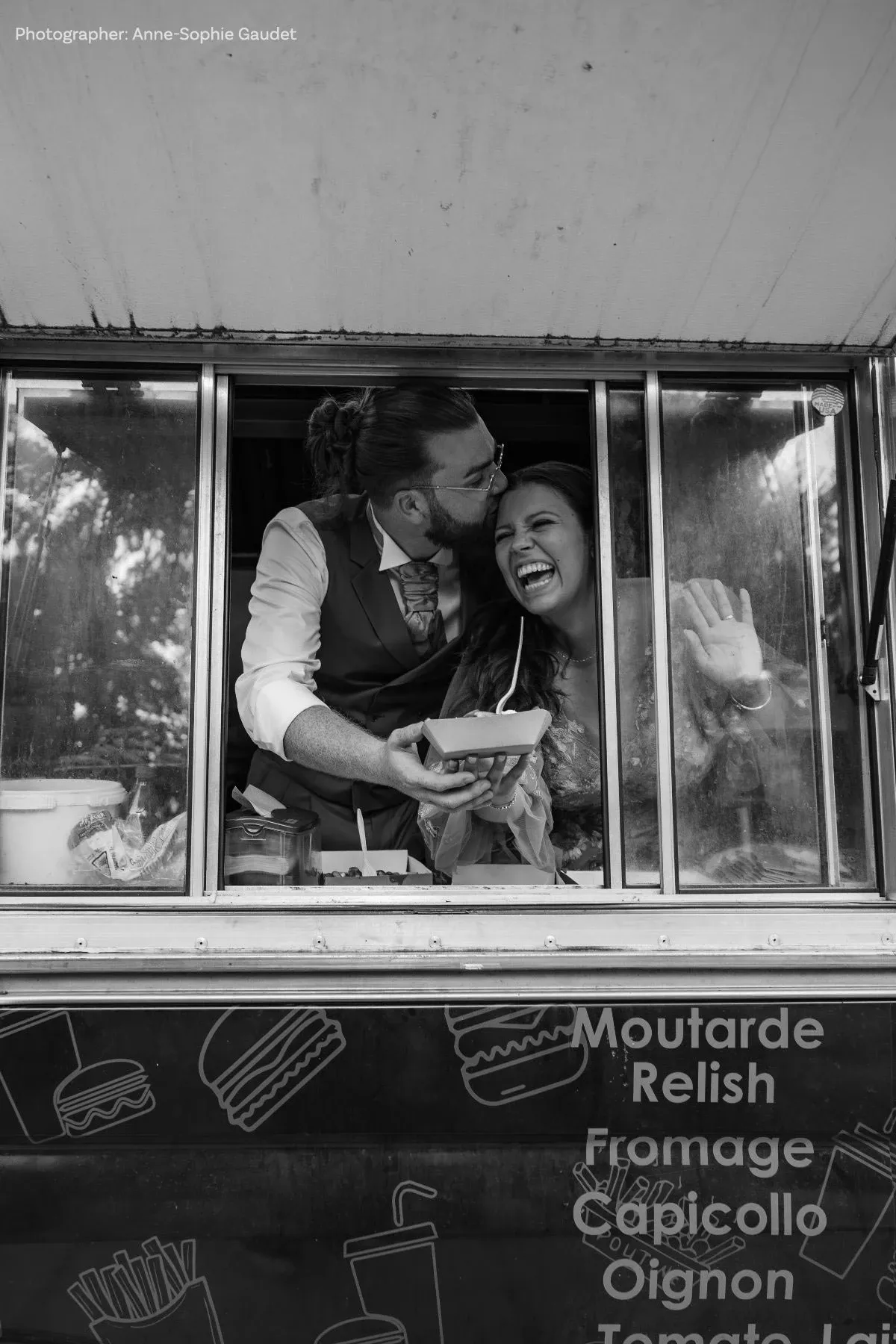 Un couple joyeux se tient à la fenêtre d'un food truck, symbolisant un moment informel mais tendre de célébration de mariage. L'homme, vêtu d'une chemise et d'un gilet, embrasse affectueusement la femme sur la tête, tandis qu'elle rit de manière éclatante, exprimant une ambiance détendue et festive. En arrière-plan, quelques éléments de cuisine sont visibles à l'intérieur du véhicule, et les mots 'Moutarde,' 'Relish,' et 'Fromage' sont inscrits sur la carrosserie extérieure, suggérant un menu de style décontracté. Cette image capture un instant spontané et heureux dans le cadre d'une réception de mariage moderne et originale.