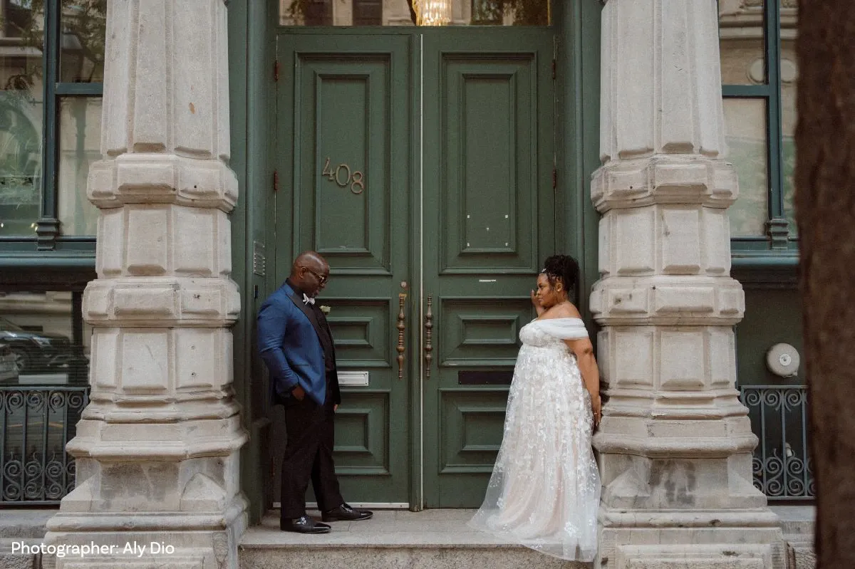 Un couple de mariés se tient sur les marches devant une élégante porte verte en bois ornée de détails architecturaux classiques. La mariée porte une robe blanche en dentelle avec des épaules dégagées, tandis que le marié est en costume bleu marine avec une chemise blanche. Ils se regardent affectueusement, créant une ambiance romantique et joyeuse. L'emplacement semble être un bâtiment historique ou une salle de réception, ajoutant une touche de sophistication à leur mariage.